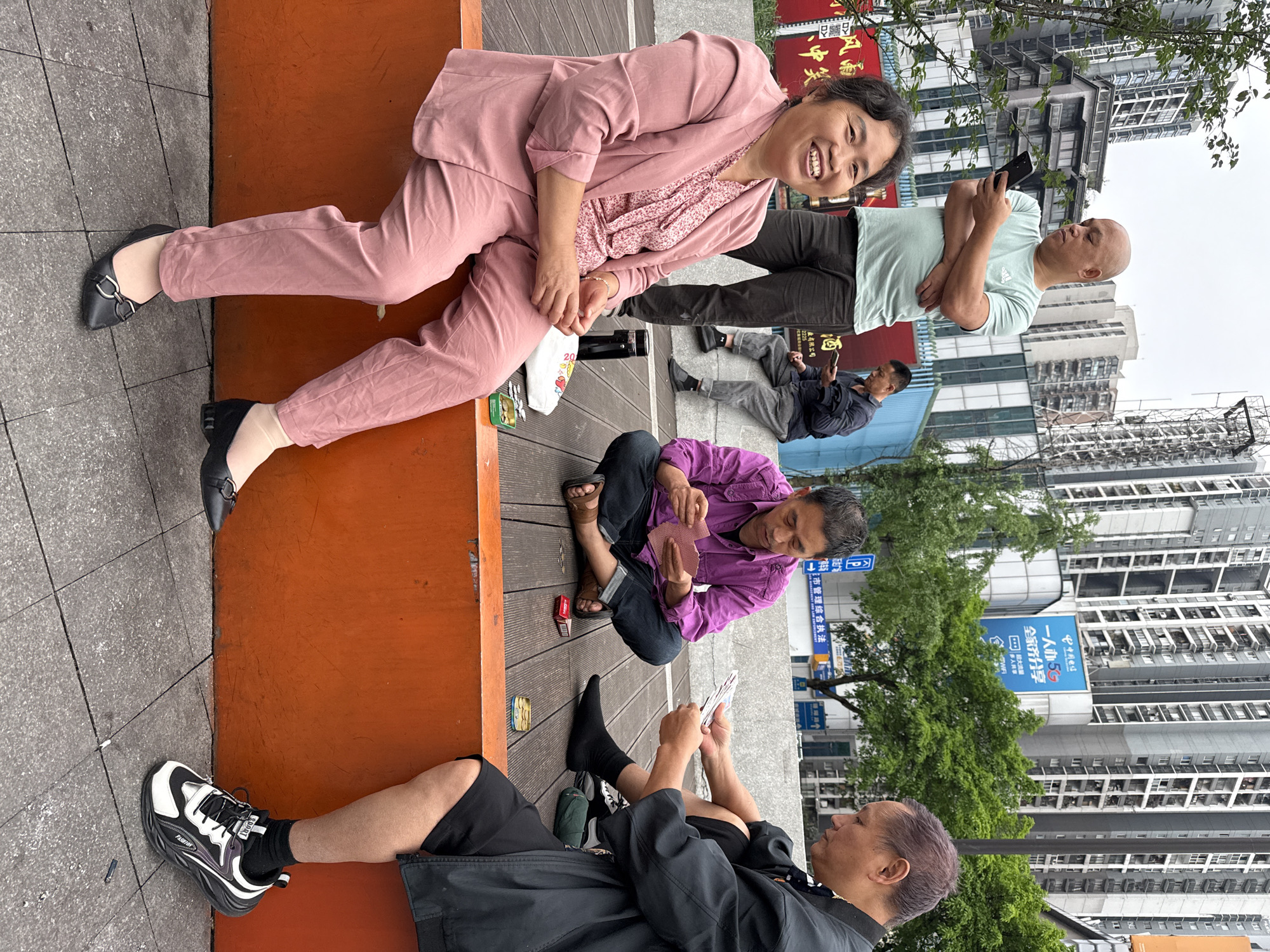 The image depicts a street scene in Nan'an District, China, with several people engaged in various activities. A woman in a pink suit is sitting on an orange bench, smiling at the camera. To her right, a bald man is looking at his phone. Another man is lying on the bench. A woman in a purple shirt is sitting on the ground playing cards, and next to her another man is playing cards, wearing black shorts, a black shirt, black socks, and a black-and-white shoe.
Buildings, trees, and street signs are visible in the background, indicating an urban environment. One of the street signs includes Chinese writing and the logo of a telecommunications company.
