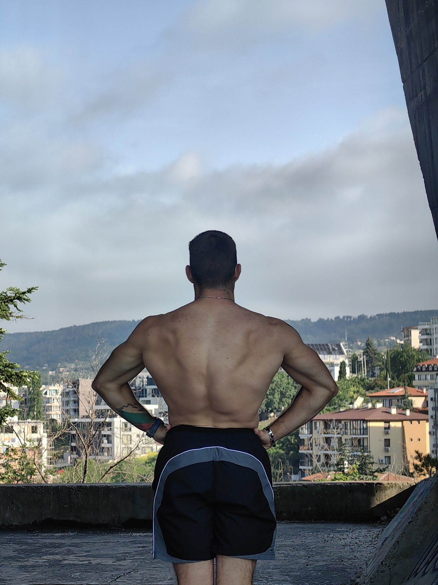 The image, taken from a rooftop in Varna, Bulgaria, captures a shirtless man from behind, his hands on his hips, displaying his toned back and arms. He wears black athletic shorts with grey and white trim. In the background, a sprawling cityscape unfolds, with buildings of various architectural styles nestled amidst lush green trees and rolling hills under a cloudy sky. A concrete structure frames the right side of the image, adding a stark contrast to the natural and urban landscape beyond. The man's tattooed arm, featuring a design in the colors of the Bulgarian flag, is partially visible.