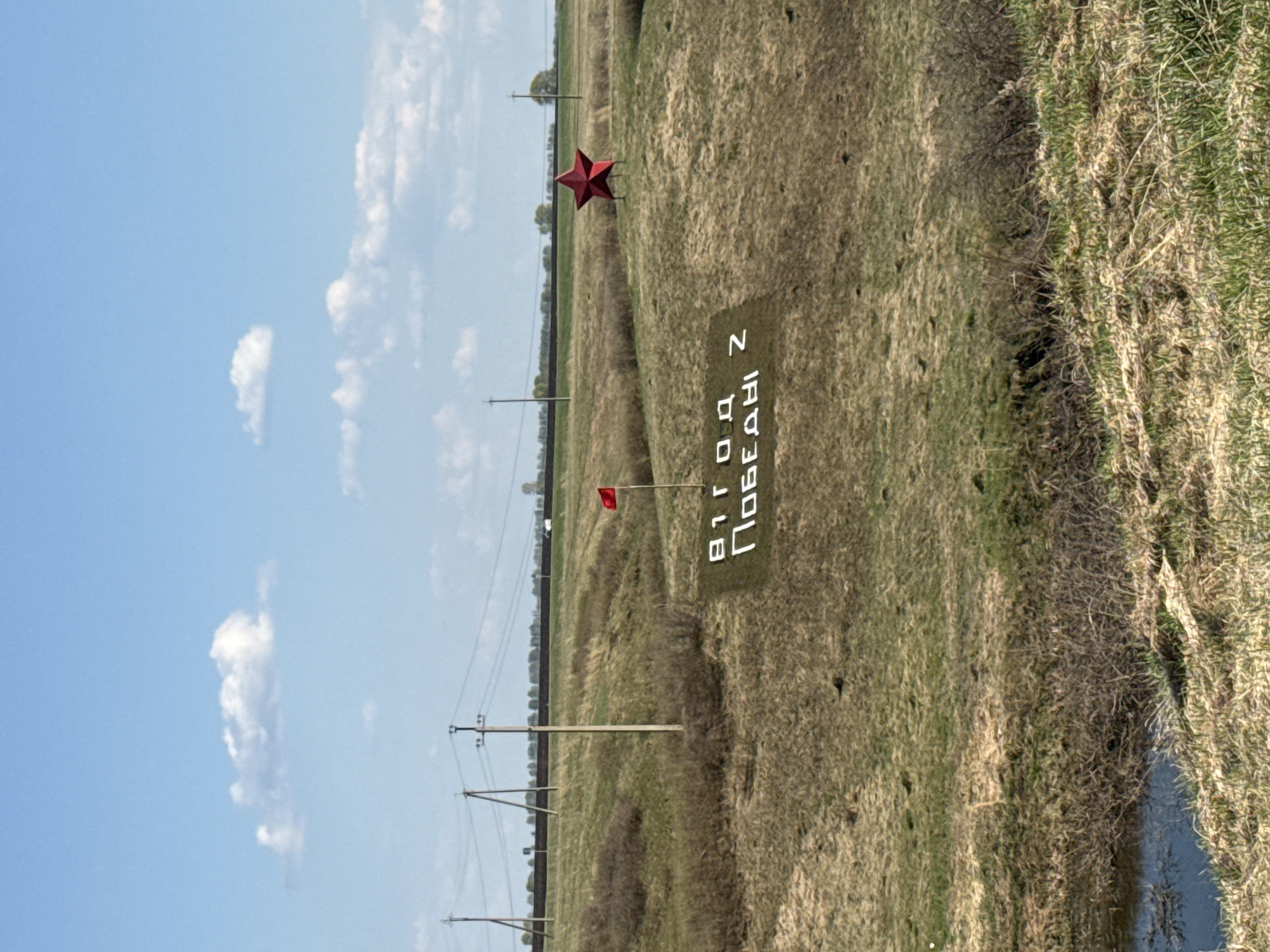 The image shows a grassy field in Такарликово, Russia. A monument commemorates the 81st anniversary of Victory Day with Cyrillic text on a sign planted in the ground. A red star is positioned further back in the field. A small red flag is situated closer to the monument. Power lines stretch across the horizon under a blue sky with scattered white clouds. A narrow stream or ditch runs along the right edge of the image.