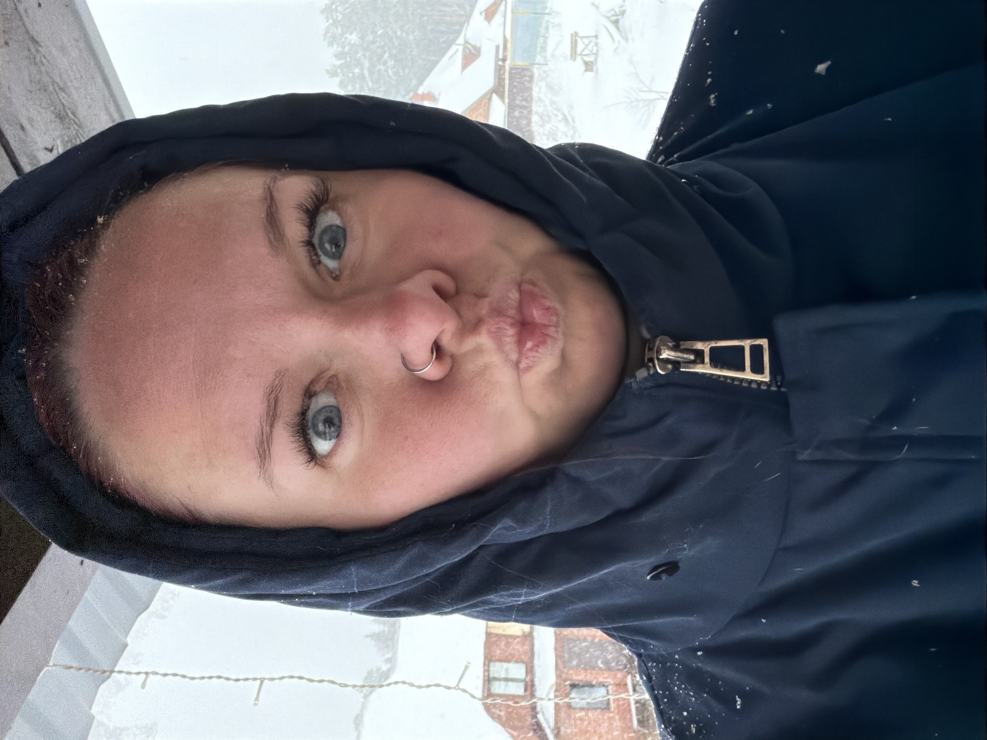 A close-up selfie depicts a fair-skinned individual with blue eyes and a nose ring, wearing a dark hooded jacket. The person is puckering their lips. The background suggests a snowy environment, with buildings and trees visible through a window, which hints to the location being Chaikovsky, Russia. The presence of snow on the jacket and visible in the background confirm a winter setting. The lighting is somewhat diffused.