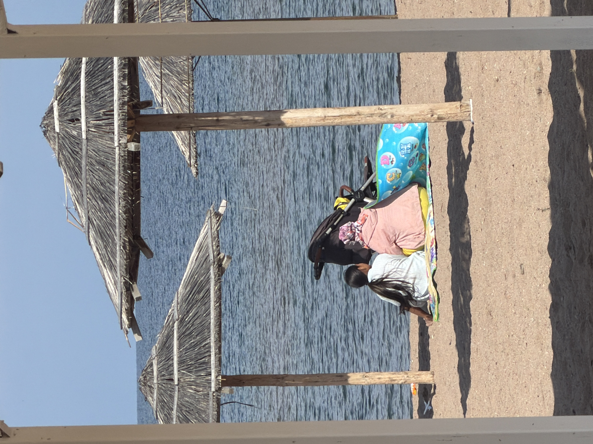 This is a medium shot of a beach scene on a clear day, as seen through a white structure. In the foreground, a sandy beach is visible with shadows cast by unseen objects. Two individuals are seated on a mat facing the water. The person on the left is a child with long dark hair. The person on the right is wearing a pink top and a headscarf. A black baby stroller is behind them, along with a colorful inflatable toy. In the middle ground, the calm blue water of the sea stretches to the horizon under a clear sky. Several rustic straw umbrellas are dotted along the beach, providing shade. The scene appears to be a public beach.