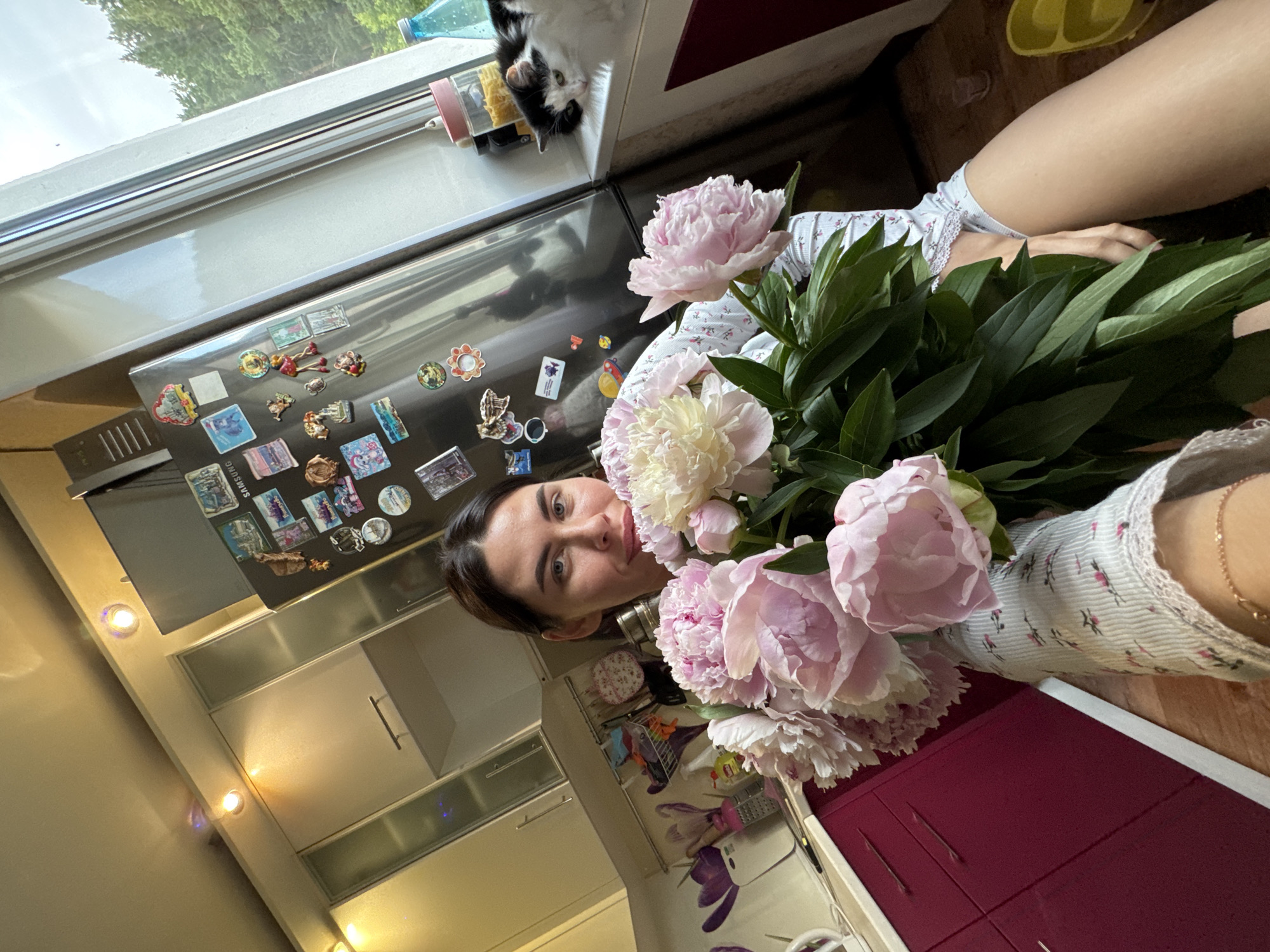 The image depicts an indoor scene, likely a kitchen. A woman is holding a large bouquet of pink and white peonies. She is wearing a light-colored shirt with a floral pattern and a bracelet on her wrist. The background shows kitchen cabinets, a refrigerator adorned with magnets, and a window with a view of trees. A black and white cat is visible on the windowsill. The overall lighting suggests daytime. The provided metadata indicates the location as Chaikovsky, Russia.