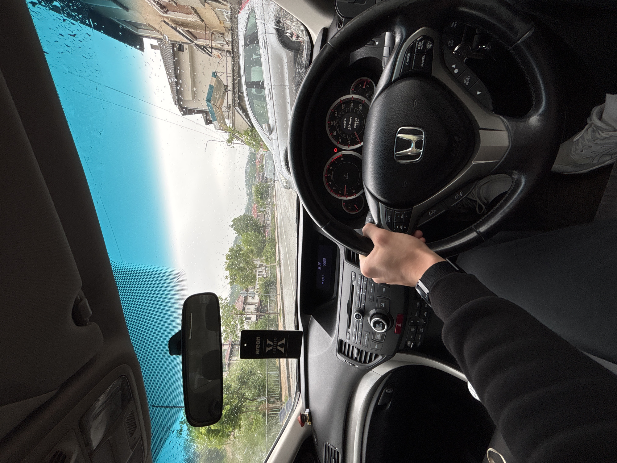The image shows the interior of a car, with the driver's hand on the steering wheel. The steering wheel has a Honda logo in the center. The dashboard is visible, with various buttons and controls. The windshield has raindrops on it, obscuring the view outside. In the background, there's a street scene with buildings and trees, reflecting the location in Zlatograd, Bulgaria. There is an air freshener hanging from the rearview mirror. The driver is wearing a watch. A parked gray car is visible through the windshield. The odometer reading on the dashboard is 158500.