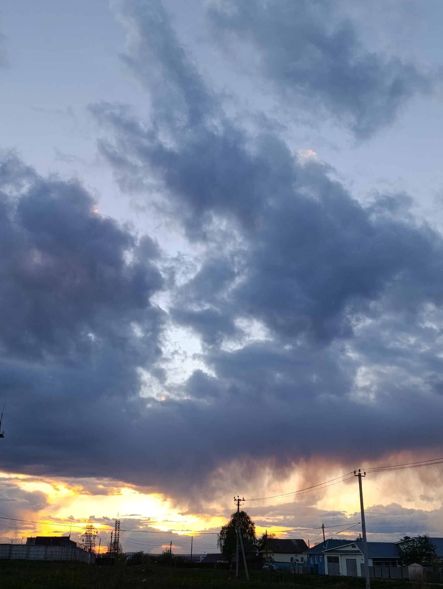 The image shows a cloudy sky over the city of Chaikovsky, Russia. At the bottom of the frame are buildings and structures, including a building with a blue roof, a small building with a green door, and telephone poles with wires running between them. Above, the sky is filled with heavy, dark clouds with some areas of yellow and orange light breaking through near the horizon, suggesting a sunset or sunrise. The overall scene conveys a sense of weather and the urban landscape.