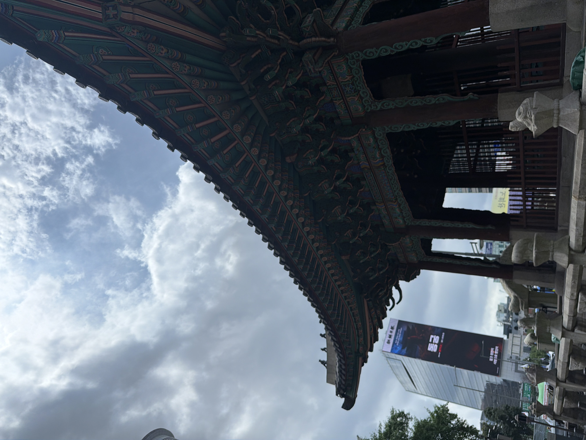 The image depicts a traditional Korean pavilion with an elaborately decorated, curved roof, set against a cloudy sky. The pavilion's eaves are adorned with intricate patterns and vibrant colors, primarily green, red, and gold. The structure is supported by dark wooden pillars. In the background, a modern, tall glass skyscraper stands prominently, featuring a large billboard advertising a Netflix production with Korean text and imagery of what appears to be a monster or creature. The billboard's presence suggests an urban setting, likely within Seoul, South Korea, given the Korean language. In the foreground, a stone balustrade with decorative elements, possibly representing animals, lines a pathway. The overall scene presents a juxtaposition of traditional Korean architecture and contemporary urban development. No people are clearly visible or are the subject of the image.