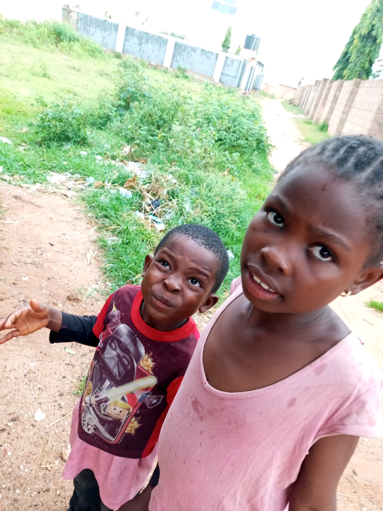 Two young children, a boy and a girl, are standing on a dirt path in what appears to be a residential area in Tempure, Nigeria. The boy, on the left, is wearing a red long-sleeved shirt with a Star Wars graphic featuring Darth Vader and a Lego character. He is looking up and to his left, with his mouth slightly open and his right hand extended. The girl, on the right, is closer to the camera and is wearing a light pink tank top. She is looking directly at the camera with a slight smile. The path is lined with dry grass, scattered debris, and some green vegetation on either side. In the background, there are fences and buildings, suggesting a semi-urban environment. The sky is overcast, indicating a cloudy day. The overall scene depicts a candid moment of two children in their environment.