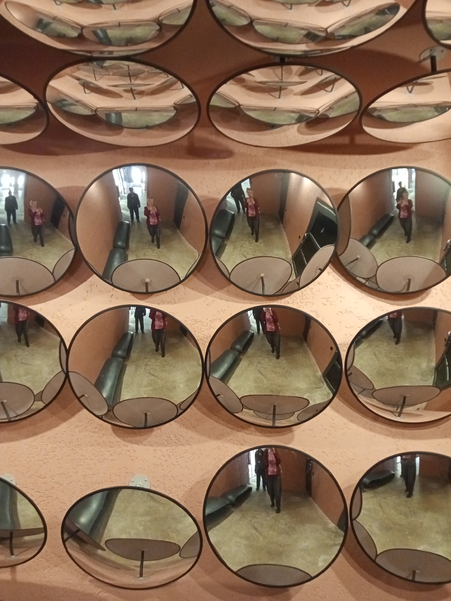 The photograph depicts a wall decorated with a grid of circular mirrors, reflecting a hallway. The mirrors are convex, distorting the reflected image. Within the reflections, two figures are visible: one in a dark coat and the other in a pink shirt. The hallway has a light-colored floor, possibly tiled, and a dark bench. The mirror installation is found on a wall in the Grand Maket Rossiya in Saint Petersburg, Russia.