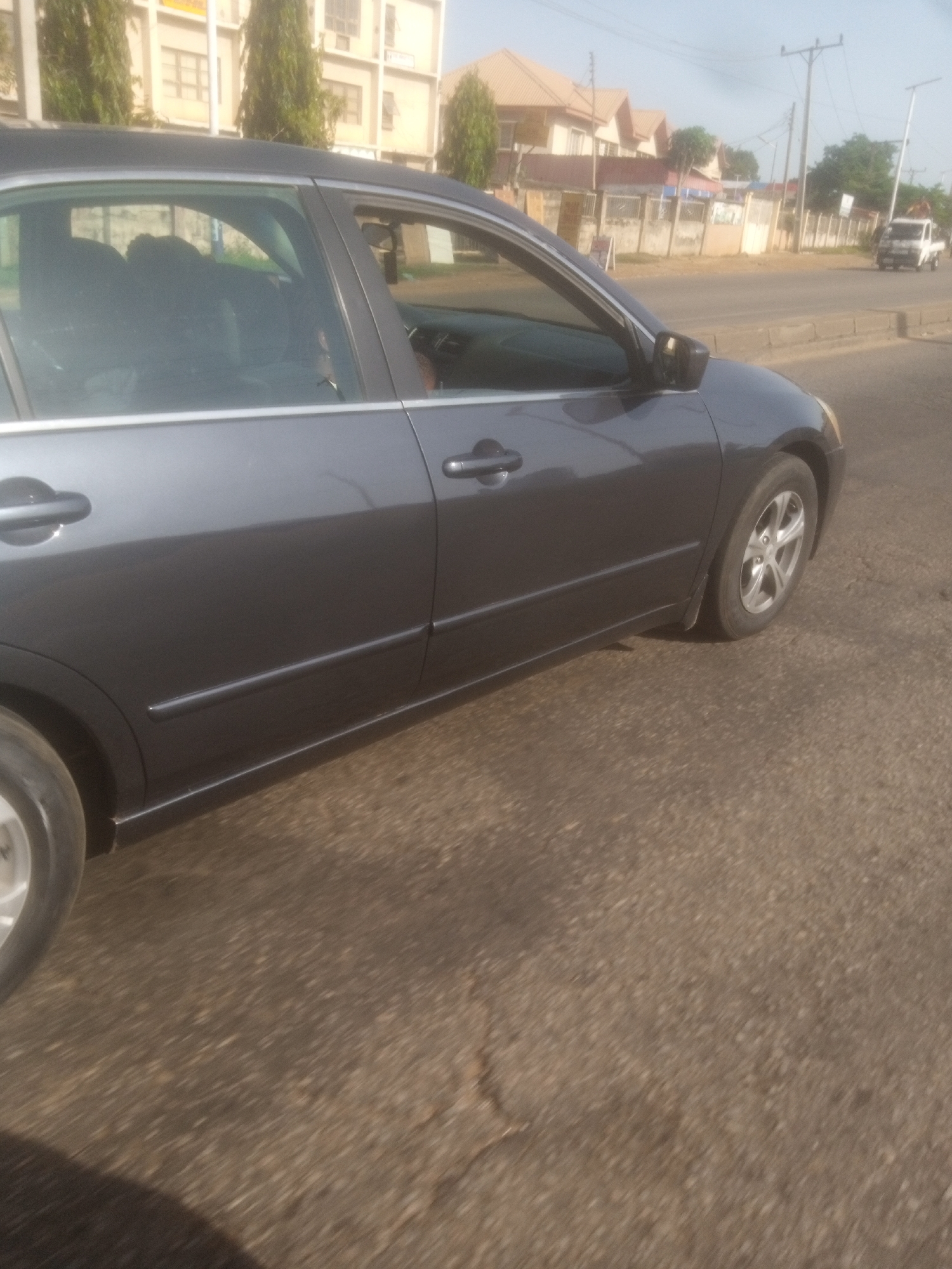 In Barnawa, Nigeria, a dark grey sedan is parked on an asphalt road. The car's left side is prominently featured, showing the front and rear doors, the side mirrors, and the wheels. The interior of the car is visible through the windows, with what appear to be two individuals sitting in the front seats. The background consists of a street with a sidewalk, buildings, trees, and utility poles. A white pickup truck is seen in the distance on the road. The lighting suggests it is daytime.