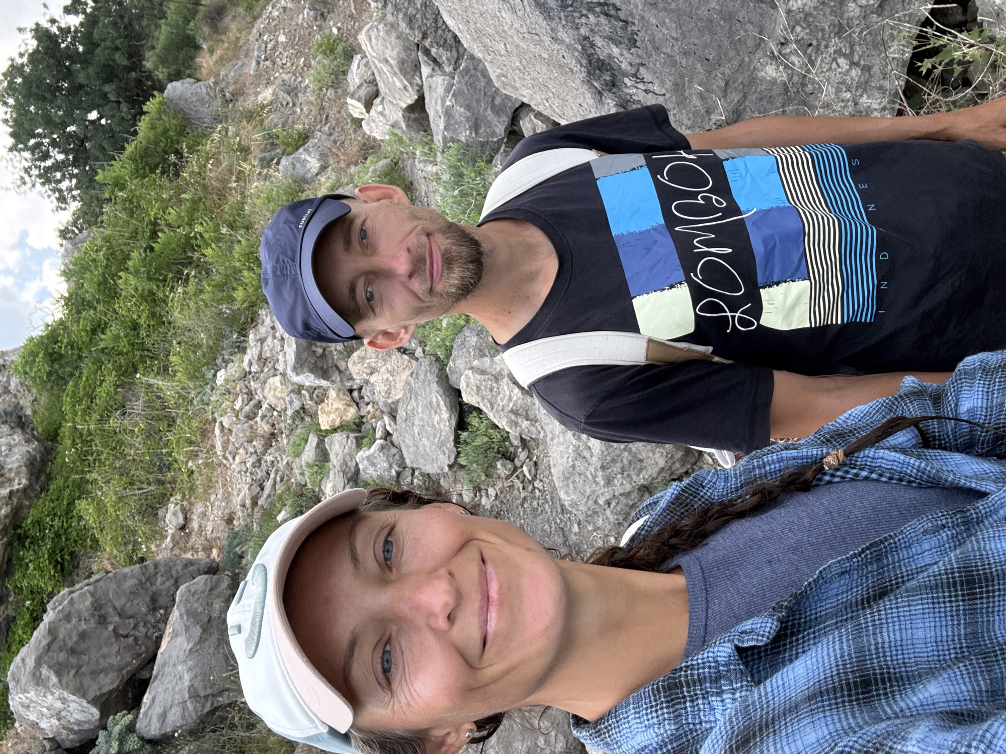 A mid-shot, outdoor selfie features a Caucasian man and woman standing amongst rocks and foliage. The woman, on the left, has a fair complexion, blue eyes, and long, dark hair braided down her back. She wears a light blue plaid shirt over a grey ribbed long-sleeved shirt and a light blue baseball cap. The man, on the right, has a fair complexion, short brown hair, and a goatee. He wears a dark blue baseball cap, a black t-shirt with a colorful graphic design, and a light-colored backpack. Both individuals are smiling at the camera. The background consists of large grey rocks and green vegetation, suggesting a natural, possibly mountainous or hilly, environment. The sky is visible in the upper portion of the image, with scattered clouds. The image does not provide enough detail to identify the specific location within Bondarenkovo, Ukraine.