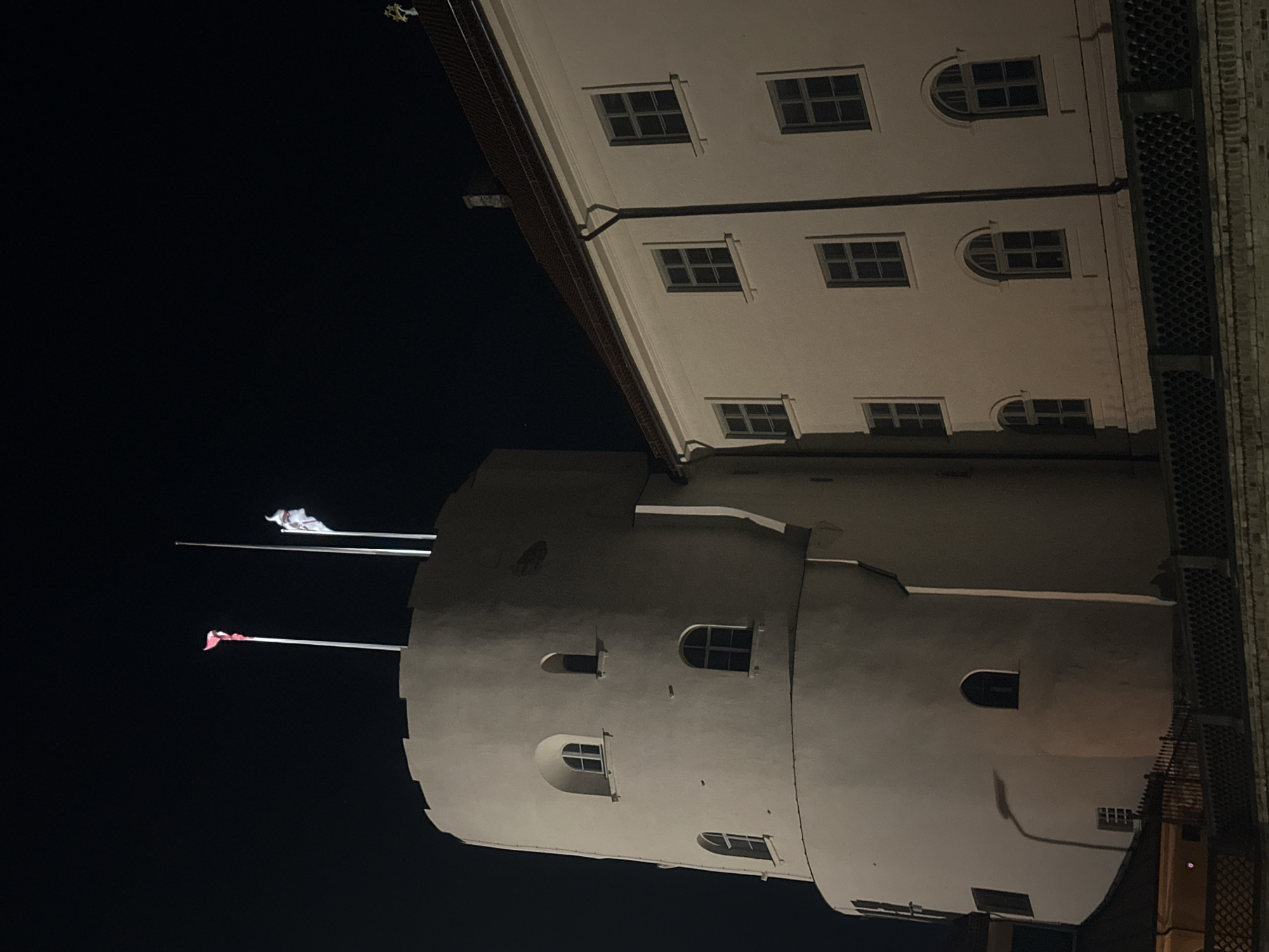 At night, a building with a cylindrical tower stands prominently in Riga, Latvia. The tower, positioned beneath the primary structure, features several arched windows of varying sizes and two flagpoles extending from its top. One flagpole displays a white flag, while the other holds a red flag. The main building has a flat façade with regularly spaced square and arched windows, exhibiting a light color. A dark sky forms the backdrop. The orientation of the image gives an unusual, tilted perspective.