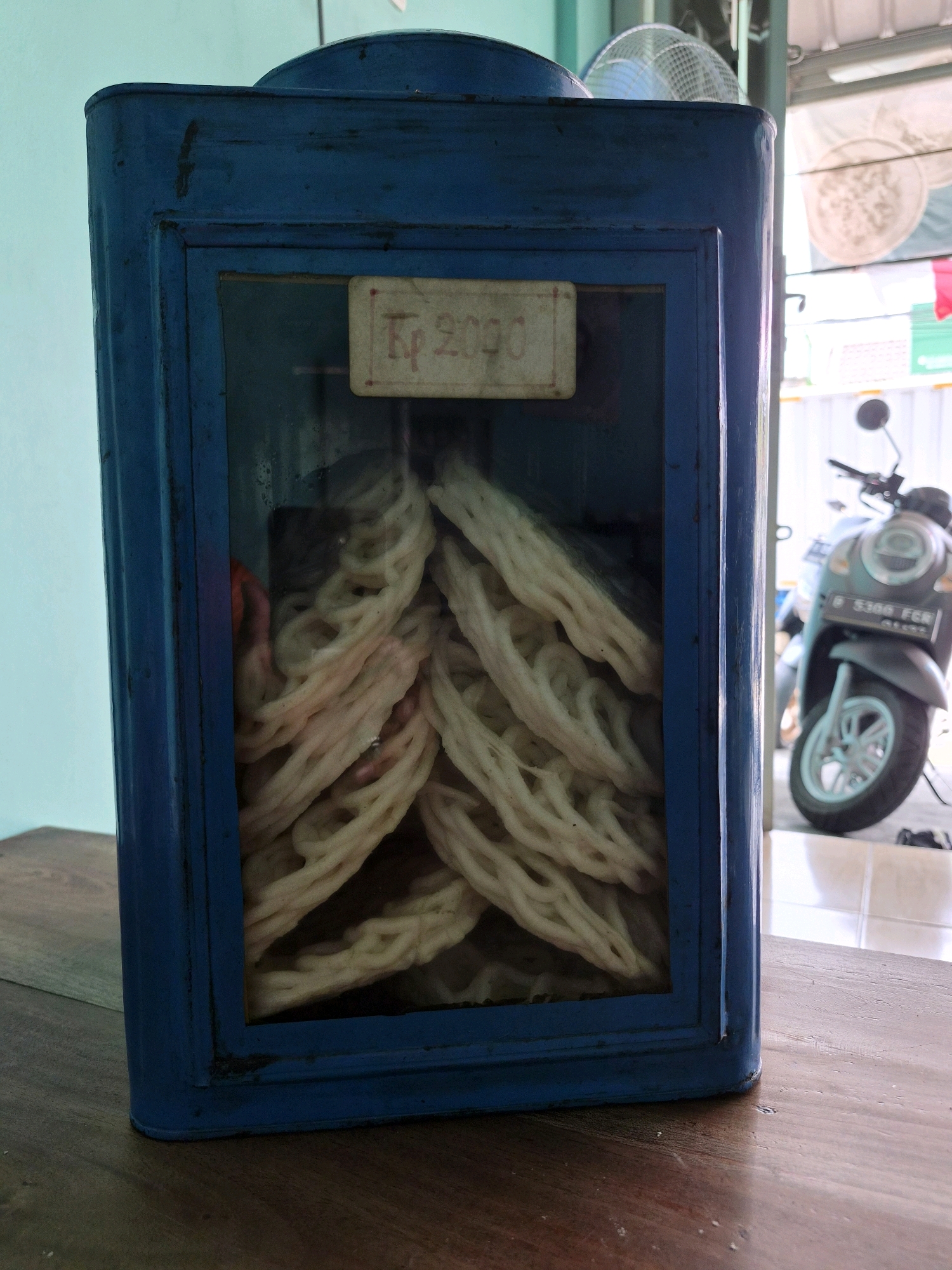 A weathered, blue, tin display case filled with white, rope-like crackers sits on a wooden table in what appears to be an outdoor stall or shop in Wanajaya, Indonesia. A small, hand-written price tag with "Rp 2000" is affixed to the glass front of the display case. Behind the case, a portion of a light blue wall is visible. To the right, a grey scooter is parked, and beyond that, a glimpse of a building with a white awning and possibly hanging laundry can be seen. The scene suggests a casual, possibly roadside vendor setting.