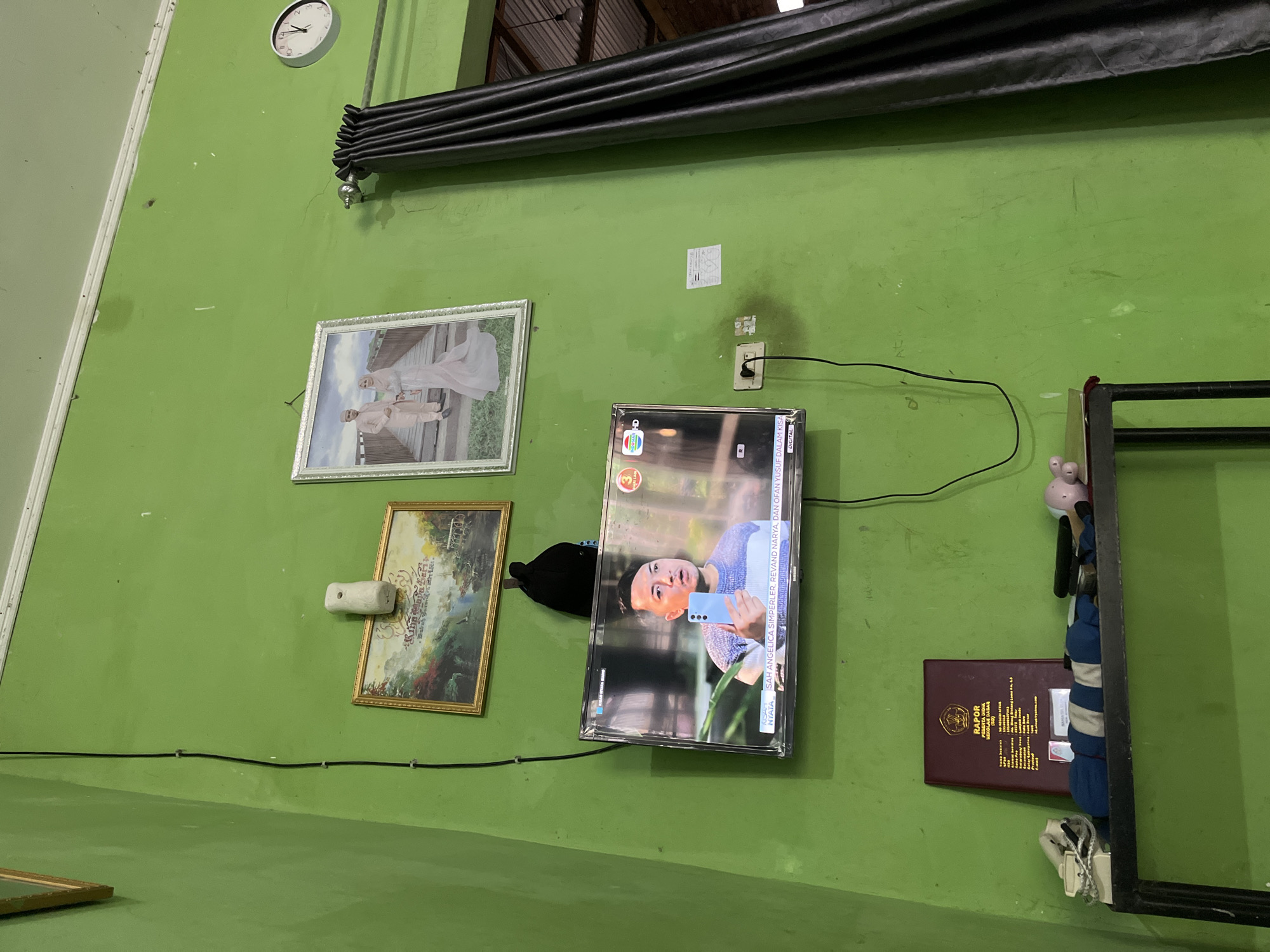 The image depicts an indoor scene, likely a living room, with vibrant green walls. A television screen displays a man holding a smartphone, with text overlays indicating a program title and names. Above the television, a framed picture shows a couple in wedding attire. To the left of that, another framed picture displays a landscape with text. A black cap hangs on the wall below the landscape picture. A white air freshener is mounted on the wall above the landscape picture. To the right of the wedding picture, a round wall clock shows the time. Dark grey curtains hang from a rod on the right side of the frame, obscuring part of the view. Below the television, a dark shelf holds various objects, including a maroon-bound report book titled "RAPOR PESERTA DIDIK SEKOLAH DASAR," a white object resembling a cordless phone handset and base, a blue and white striped fabric, and what appear to be children's toys. A black electrical cord snakes down the wall from the television to an outlet. The overall impression is of a lived-in space with personal mementos and everyday electronics. There is no information in the image to suggest the location is the city of Babahjurong, Indonesia.
