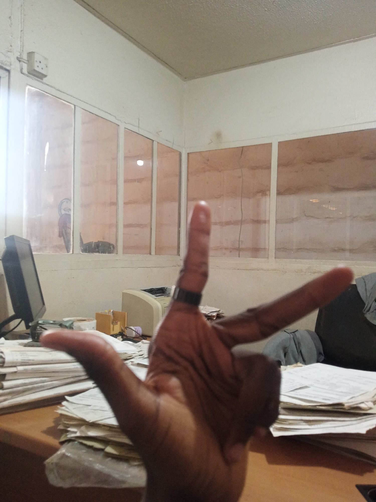 The image captures an indoor scene, likely an office in Kakuri Gwari, Nigeria. In the immediate foreground, a person's hand with dark skin is raised, fingers spread in a gesture that might represent the number three, an "M" or "W" sign, or simply a pose for the camera. A dark ring is visible on the middle finger.
The background reveals a cluttered office desk made of light brown wood. On the left side of the desk, an older model computer monitor with a dark casing is visible. To the right, a beige printer or fax machine sits on the desk. Numerous stacks of papers, some seemingly wrapped in plastic, are spread across the desk surface, indicating an active workspace. Other small items like a roll of tape, a cardboard box, and a spool of purple thread are also discernible on the desk.
Behind the desk, a wall features a series of framed, rectangular panels, possibly windows or translucent room dividers, through which indistinct shapes are visible, including what looks like a kettle or pot on the left. The panels appear to have a slightly textured or wavy pattern, perhaps reflecting something behind them or being made of a patterned material. The walls themselves are a light, off-white or cream color. In the upper left corner of the image, near the ceiling, a white electrical wall socket with three square pins is mounted, a common design in Nigeria. The ceiling is plain and light-colored. A dark object, possibly a bag or folded clothing, rests on a chair or surface behind the desk on the right side. No discernible text is clearly visible in the image. The overall impression is that of a functional, somewhat aged office environment.