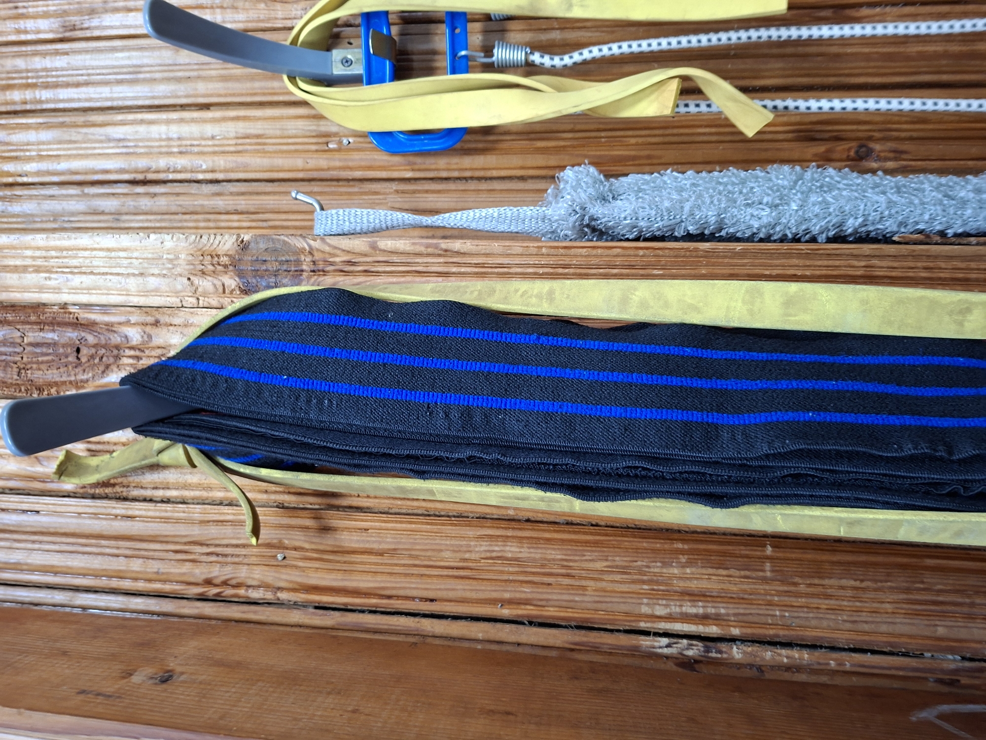 The image shows a close-up of several items hanging on a wooden wall. From left to right, there is a black fabric with blue stripes, a light gray fuzzy object, and a yellow strap with a coiled white and black cord. The items are hanging on hooks mounted to the wall. The wall is made of vertical wooden planks. This scene does not contain any information that would allow for a specific location within the city of Kazan, Russia, to be identified.