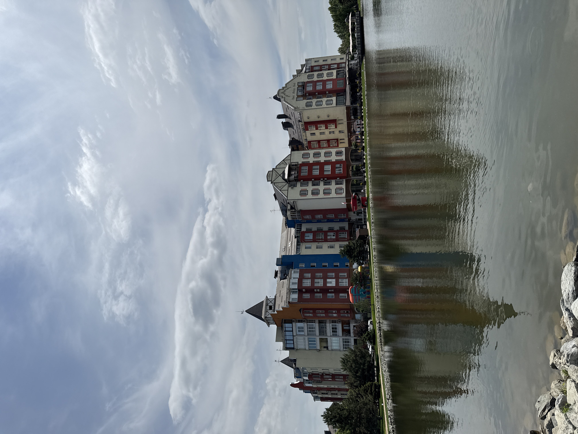 The image depicts a waterfront view of a row of colorful buildings with a lake in the foreground. The buildings feature a variety of architectural styles, with multiple stories, balconies, and pitched roofs. They are painted in vibrant colors such as red, orange, yellow, and blue, interspersed with white and beige sections. Some buildings have decorative gables and turrets.
The lake reflects the buildings and the sky, creating a symmetrical effect. The water appears calm with gentle ripples. The shoreline in front of the buildings is lined with trees and a paved walkway, with some people visible strolling along it. There are also some playground-like structures in front of the buildings.
The sky is partly cloudy, with a mix of white and gray clouds against a light blue background. The overall scene suggests a pleasant, possibly resort-like, environment.
This image is identified as the "Hotel Muller-Bach" located in the city of Krasnodar, Russia.
