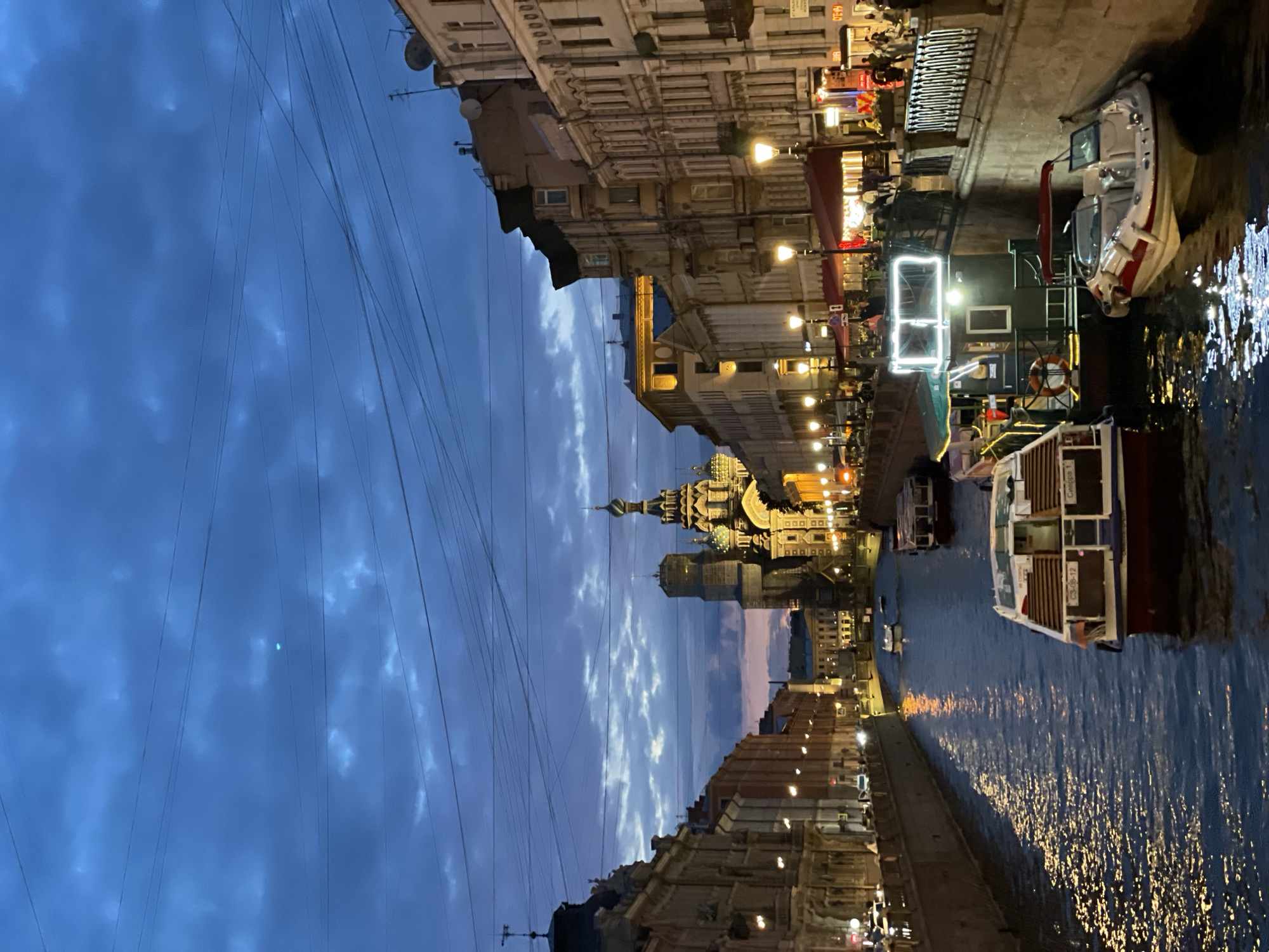 This is a full shot, taken at dusk, of a canal lined with buildings and boats. The sky is a deep, moody blue with scattered, dark clouds. A prominent church with onion domes and intricate architecture, lit by golden lights, stands at the end of the canal, partially obscured by scaffolding. The buildings lining the canal are a mix of architectural styles, with ornate facades and many windows. Streetlights cast a warm glow on the buildings and the water. Several boats are moored in the canal, including tour boats and smaller vessels. People are visible on the sidewalks and on the decks of some of the boats, some appearing to be tourists. The water reflects the lights from the buildings and boats, creating a shimmering effect. Overhead, power lines crisscross the sky. The overall scene evokes a sense of historic European charm and nocturnal city life.