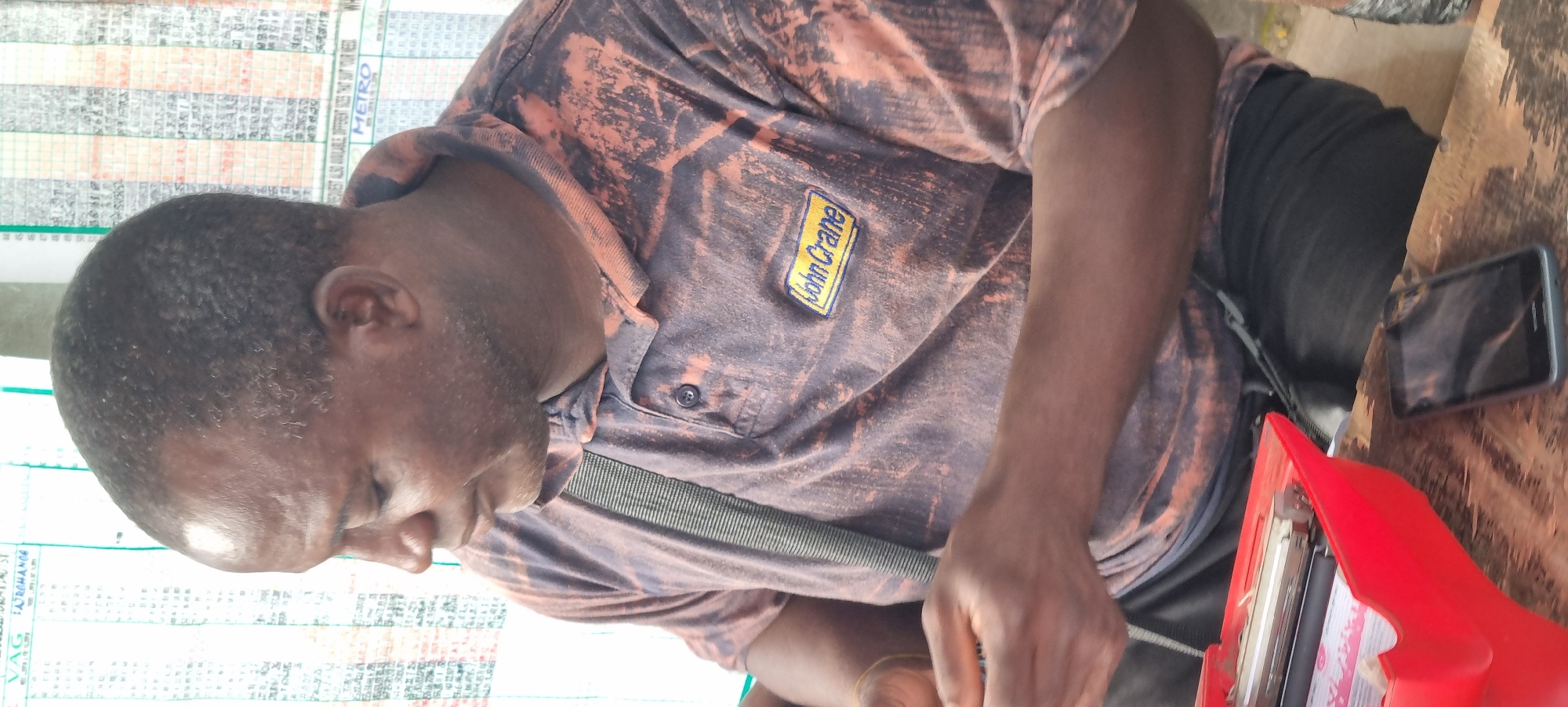 The image depicts a man focused on a task, likely related to printing or writing. He is wearing a mottled orange and black shirt with a "John Crane" logo on the chest, and he has a strap across his shoulder. To his right, there's a smartphone and a red printing or stamping device on a wooden surface. To his left is a printed sheet on a wall containing information such as "VAG AROHANCE" and "METRO". Based on the text "REQUEST ALSO AVAILABLE, DIFFERENT SITES THAT SUT YOUR NEED", it's highly plausible the setting is Lagos, Nigeria, where betting shops and lottery businesses are common. The man's activity suggests he may be involved in printing tickets or forms, and the overall environment implies a local, possibly outdoor, business setting.