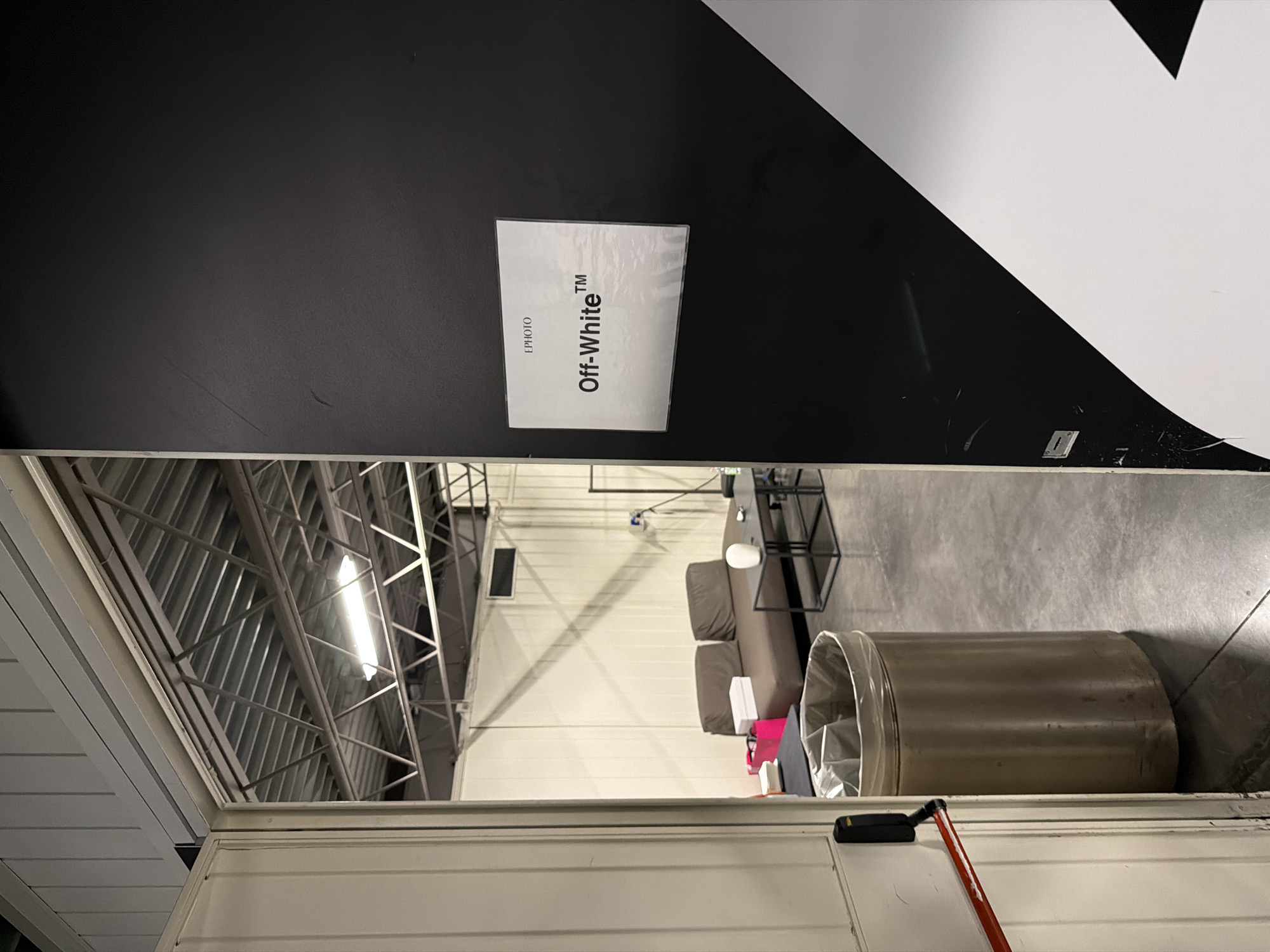 The photograph shows an interior space with industrial and design elements. A sign with the text "Off-White™" is attached to a dark-colored wall with white graphic elements. The interior space is likely a showroom or office area, as there is a couch, a small table with items, and an industrial-style ceiling with metal supports and a fluorescent light. A large cylindrical container is also visible. The image seems to be taken from inside a structure with a metal frame, looking into the interior space. The location is not specified.