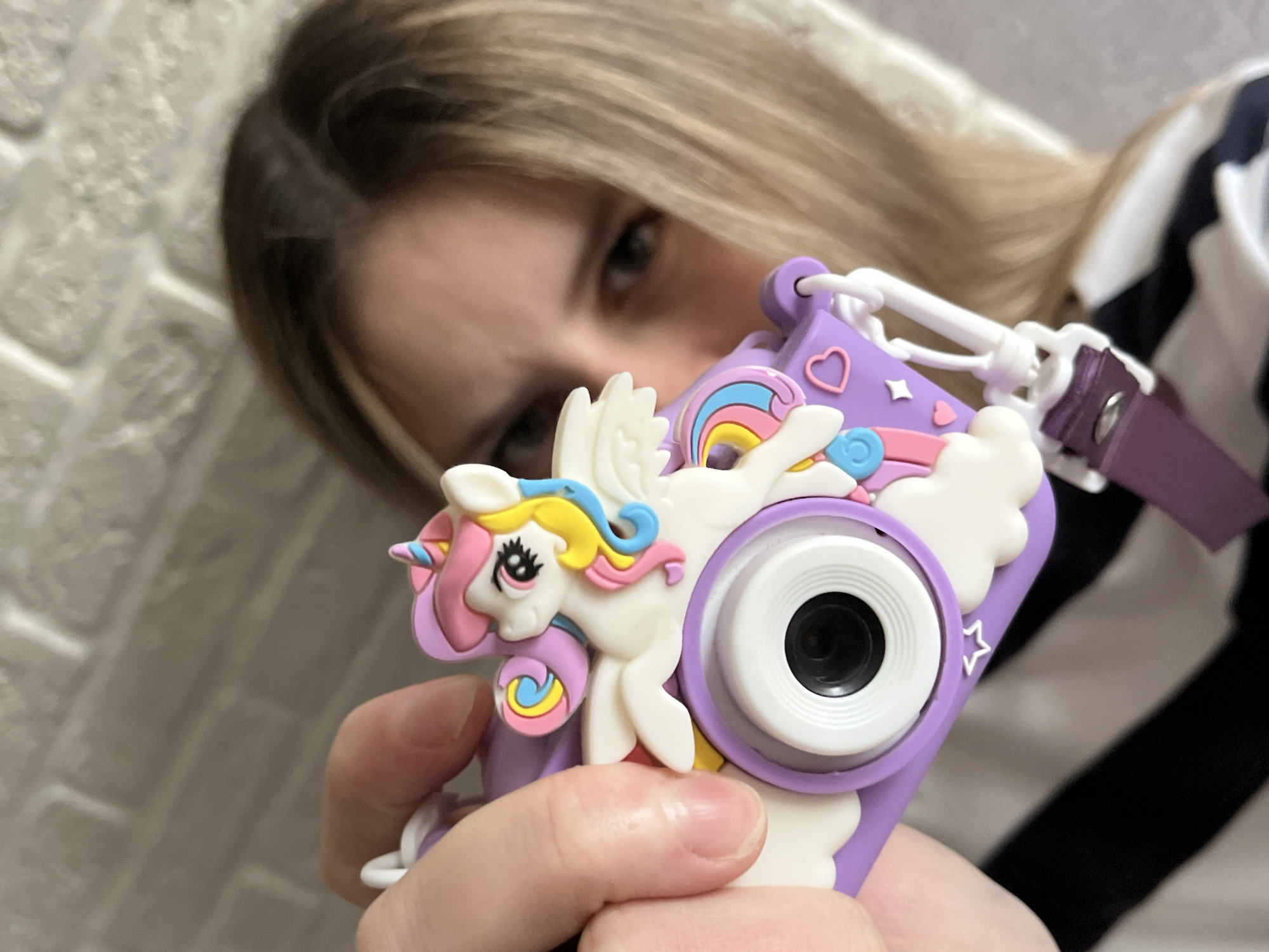 A young person with blonde hair holds a lilac-colored children's camera, decorated with a white unicorn. The camera is the primary focus, with the person's eyes visible behind it. The background includes a white brick wall on the left and a striped shirt on the right. The photograph was taken indoors, possibly in the city of Chaikovsky, Russia.