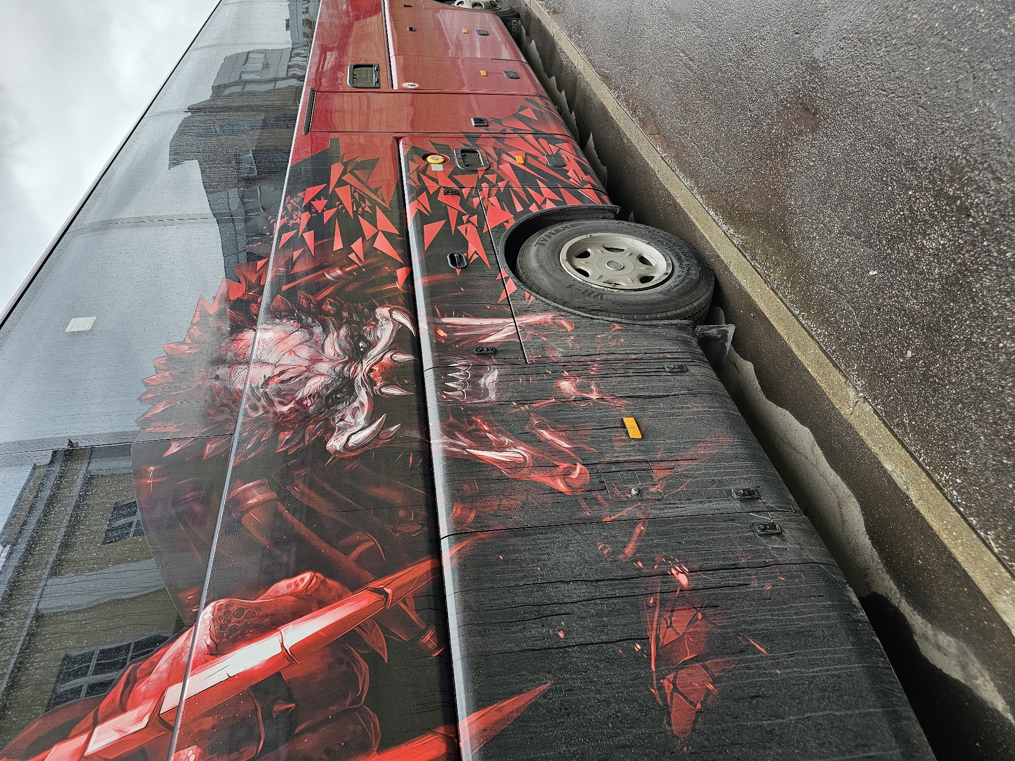 A red bus with an aggressive, red and black graphic of a monster or dragon's head is parked near a curb in what appears to be the city of Khimki, Russia. The graphic covers a large portion of the bus's side panel. The bus is positioned close to a building with a mirrored surface, reflecting the surrounding urban environment. The ground and surfaces of the bus are wet, possibly from rain. The bus's wheel is visible, situated just above the curb.