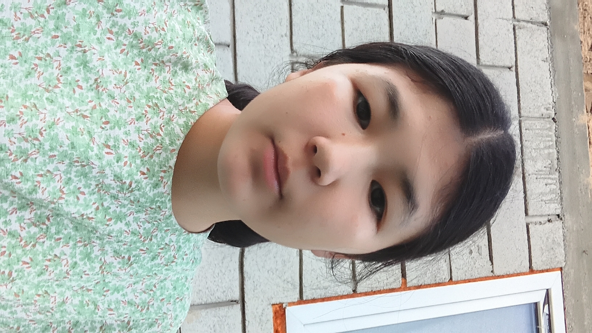 A young Asian female is photographed from the chest up, facing the camera directly. She has dark hair pulled back, with a few strands loose around her face. Her eyes are dark and wide, and her mouth is closed. She is wearing a light green t-shirt with a white and red floral pattern. The background consists of a light grey brick wall. To the right of the woman is a portion of a white window frame with an orange trim. Above the brick wall, a strip of reddish-brown construction material is visible. The scene appears to be outdoors, possibly near a building under construction. The location is identified as Uchkun, Kyrgyzstan.