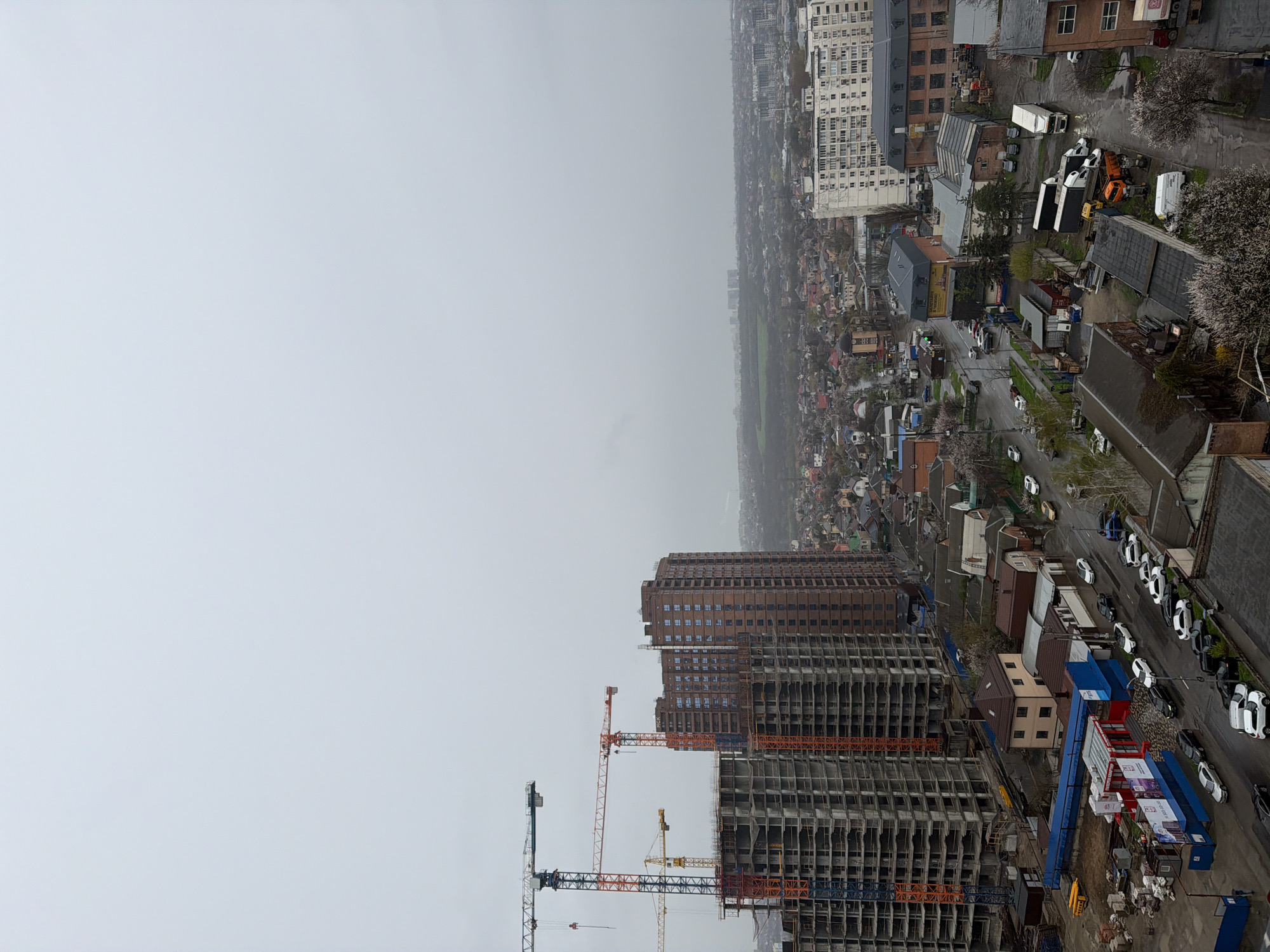 The image depicts a cityscape in Rostov-on-Don, Russia, from an elevated perspective, likely a high-rise building. The scene is overcast, with a flat, gray sky dominating much of the frame. In the foreground are high-rise buildings under construction. Construction cranes are in view, indicating ongoing development. In the midground, there is a mix of residential areas, with closely packed houses. A row of parked cars is visible along a street. In the background is a dense urban landscape.