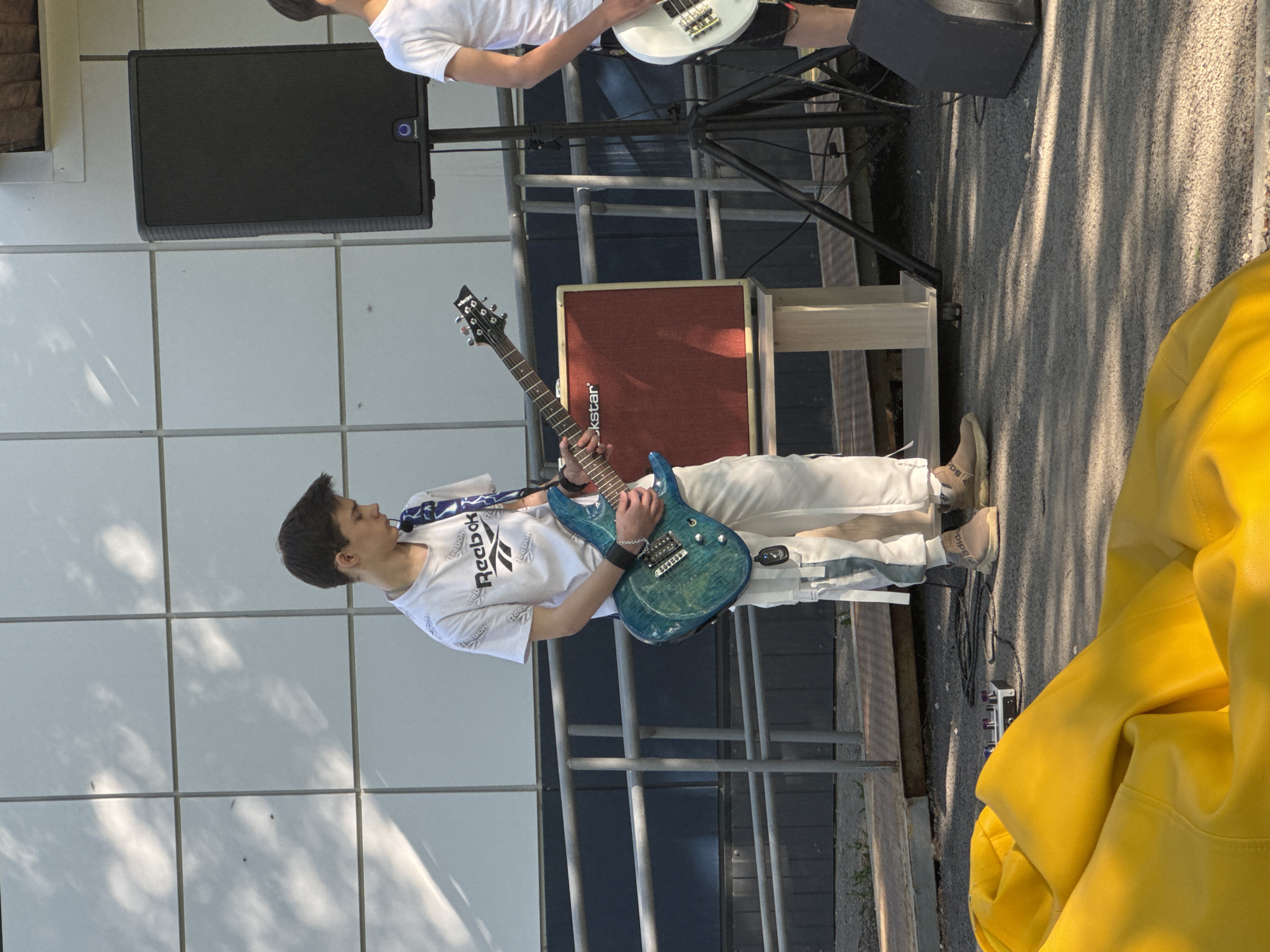 The image shows a teenager playing an electric guitar. He is wearing a white t-shirt, white pants, and beige sneakers. The guitar is blue. Behind him is a red amplifier labeled "Rockstar" and a black speaker on a stand. Another person, partially visible at the top of the frame, is playing a white bass guitar. They are on a paved surface with a metal railing. There is a large yellow object partially in the foreground on the right side. The background consists of a white wall with grid-like patterns. There is no way to determine the location in the city of Berezovka, Russia, based on the image alone.