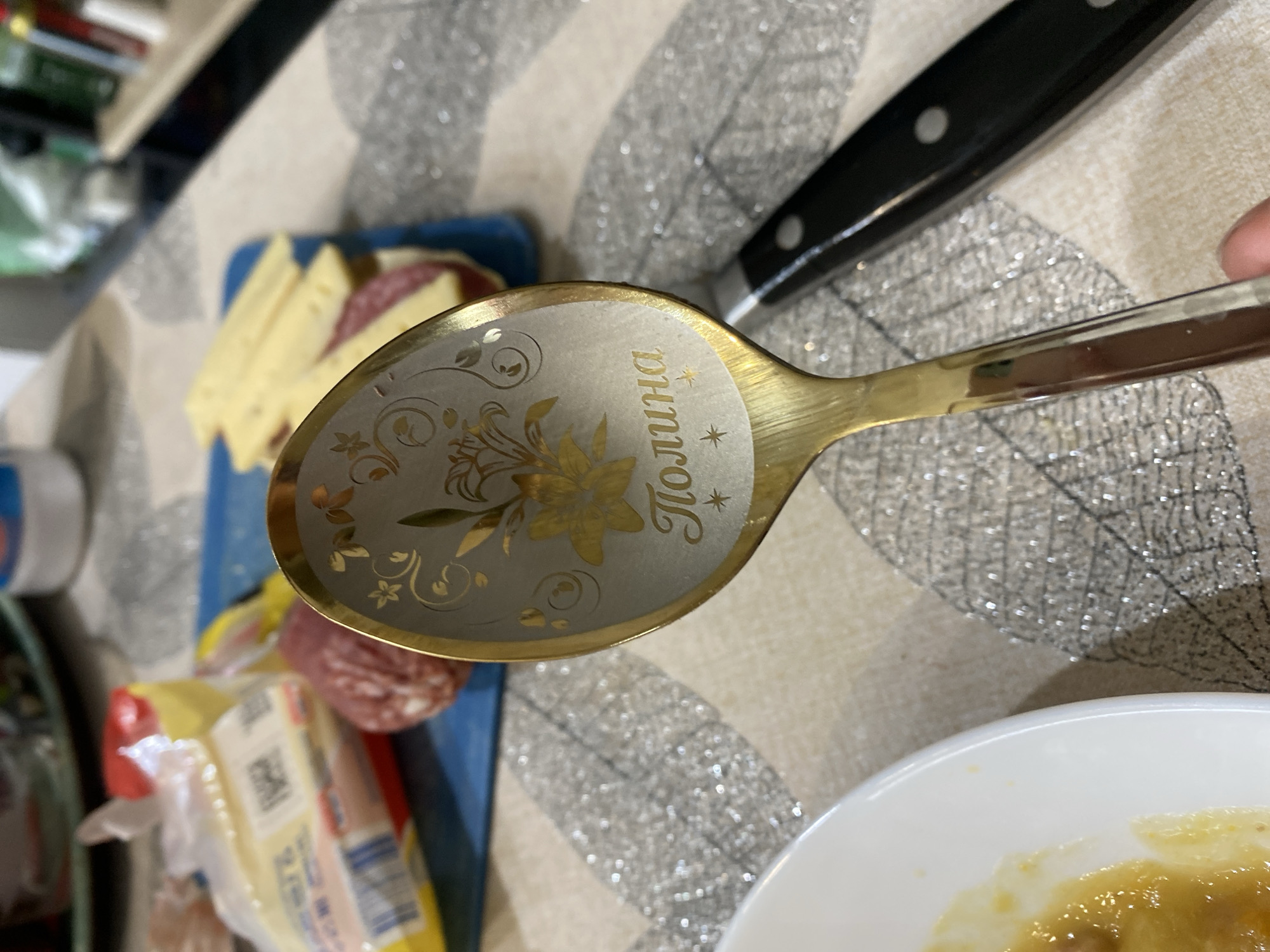 A close-up shot shows a golden spoon resting on a table, with its bowl facing the viewer. The spoon is engraved with the name "Полина" (Polina in Russian) and a floral design. In the background, partially visible, is a blue cutting board with cheese and sausage slices, and a knife. A white plate with some food is visible in the lower right corner. The setting seems to be indoors, possibly a kitchen or dining area, as suggested by the food and tableware. The specific location or city is not identifiable from the image.