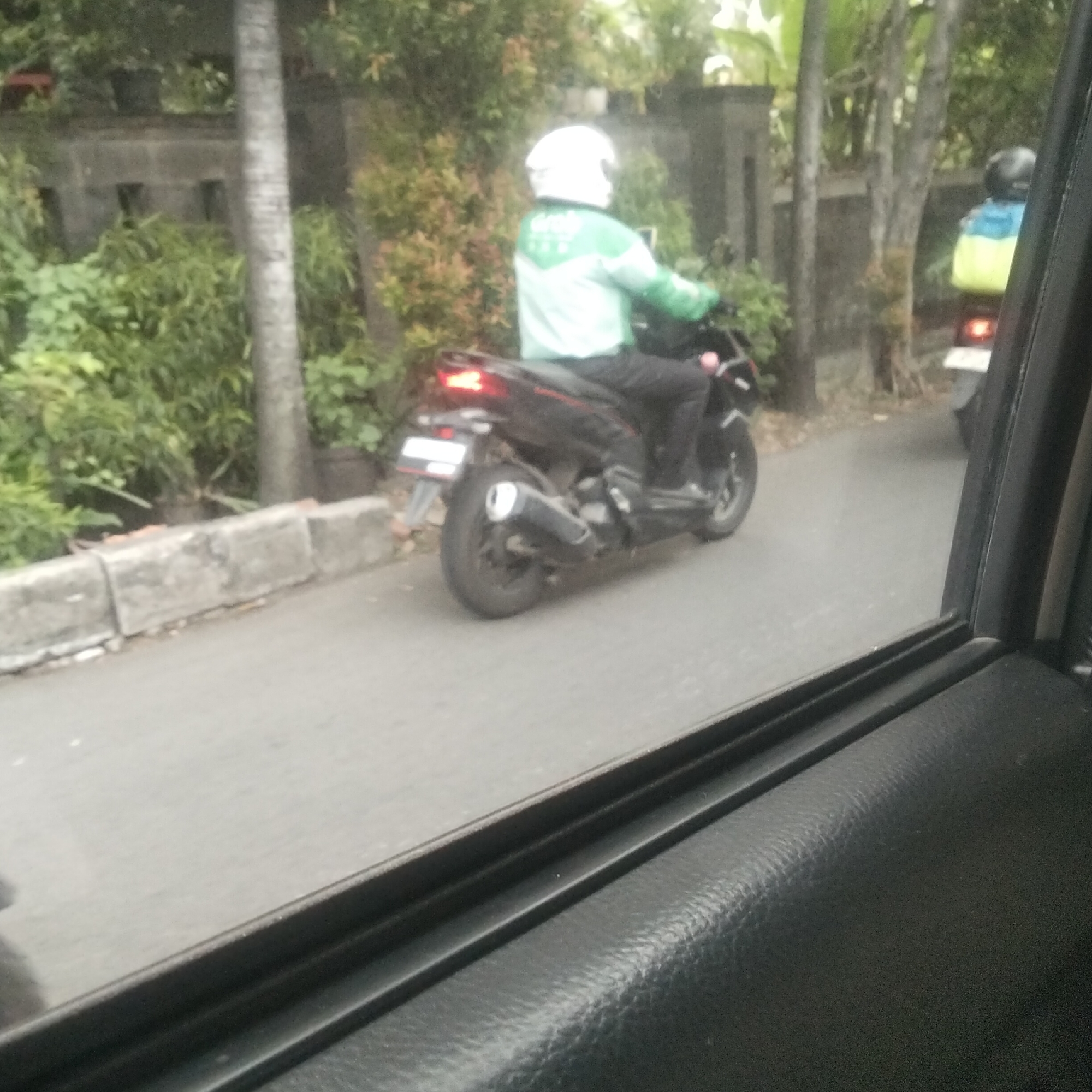 The image captures a street scene, likely in Bogor, Indonesia, viewed from inside a vehicle. A Grab delivery rider, identifiable by their green and white uniform and white helmet, is riding a black scooter. The scooter has a white license plate and a prominent exhaust pipe. In the background, lush green foliage and trees are visible, interspersed with what appears to be a low stone wall. Another scooter, ridden by a person wearing a blue and yellow jacket and a dark helmet, is partially visible further down the road. The overall impression is of a typical urban street with moderate vegetation.