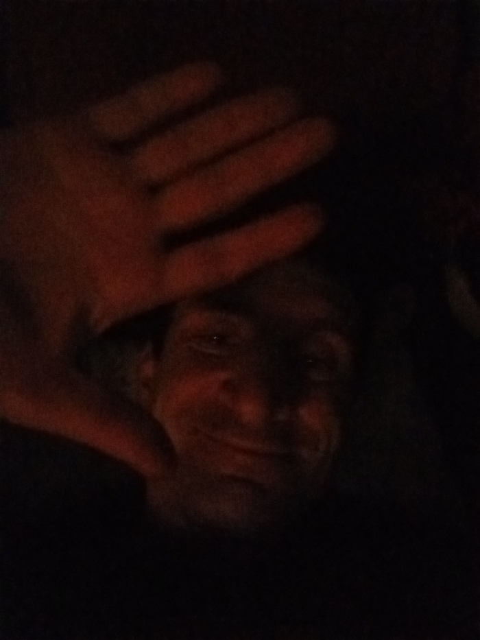 The scene depicts an individual in Yarun, Ukraine, captured under extremely low-light conditions. A human left hand is positioned prominently in the upper left quadrant of the frame, fingers slightly splayed and extended, partially obscuring the subject's face. Behind the hand, a human face is partially visible, displaying a subtle smile or smirk and showing the left eye with a faint reflection. Both the hand and the visible facial features exhibit a reddish hue, indicative of the dim lighting. The background is uniformly dark and devoid of any discernible objects or environmental features. The individual appears to be holding their hand up, partially concealing their face from the camera.