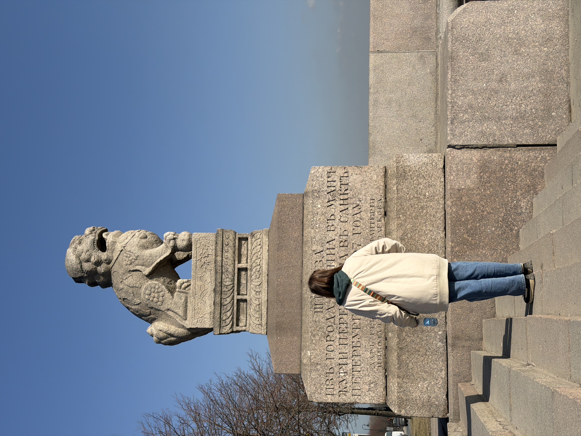 An outdoor scene captures a person standing on stone steps, facing a large stone Chinese guardian lion (Shih Tzu) statue mounted on a multi-tiered granite pedestal. The statue depicts a mythical lion with an open mouth, facing right. The pedestal bears Cyrillic inscriptions, including "ИЗЪ ГОРОДА ЖУРИ ПЕРЕ ВЪ САНКТЬ ПЕТЕРБУРГ 7 ГОДУ", which identifies the location as the Petrovskaya Embankment in Saint Petersburg, Russia, referring to the gift from Jilin in 1907.
The person is viewed from behind, wearing a beige bomber-style jacket over a dark green hoodie, blue jeans, and dark flat shoes. A multi-colored woven strap bag is slung over their right shoulder. Their left hand holds a clear plastic water bottle. They are standing still, observing the monument.
The sky is clear and bright blue, indicating fair weather. To the left, a deciduous tree with bare branches is visible. In the distant left background, a faint street or embankment road with a white vehicle and a lamppost can be discerned. The right background features a hazy horizon. The immediate foreground and monument are constructed from coarse-grained granite.
