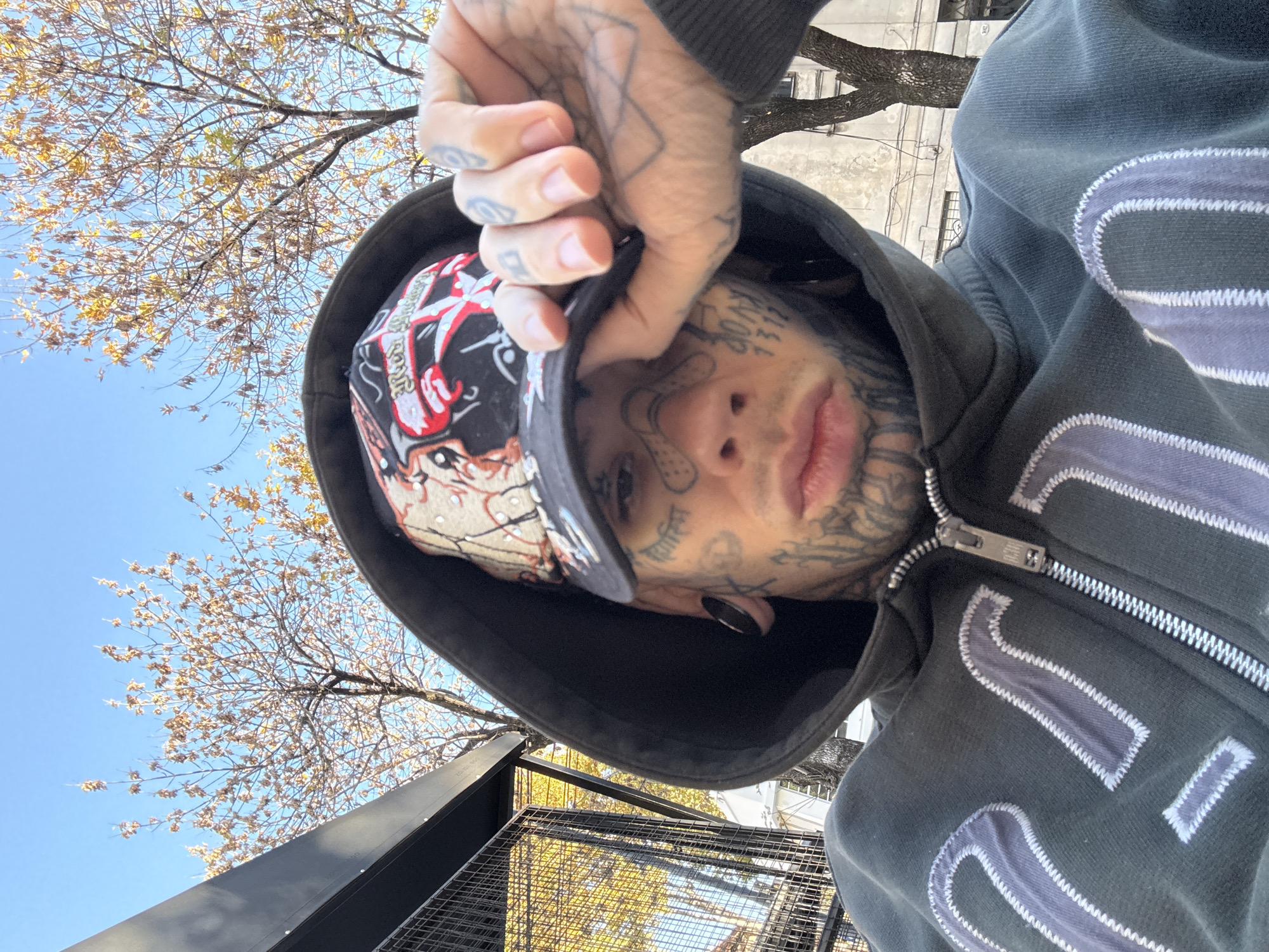 A close-up, low-angle shot depicts a person with extensive facial and hand tattoos. They are wearing a hooded grey jacket and a patterned baseball cap, partially obscured by their hand holding the brim. The jacket has large stylized numbers appliquéd on the front. The person appears to be looking directly at the camera. Above and behind them, a tree with autumnal foliage is visible against a blue sky. The background suggests an urban setting. The location is likely Buenos Aires, Argentina, though specific landmarks are not visible. The person appears to be standing or sitting near a metal structure, possibly a fence or railing.