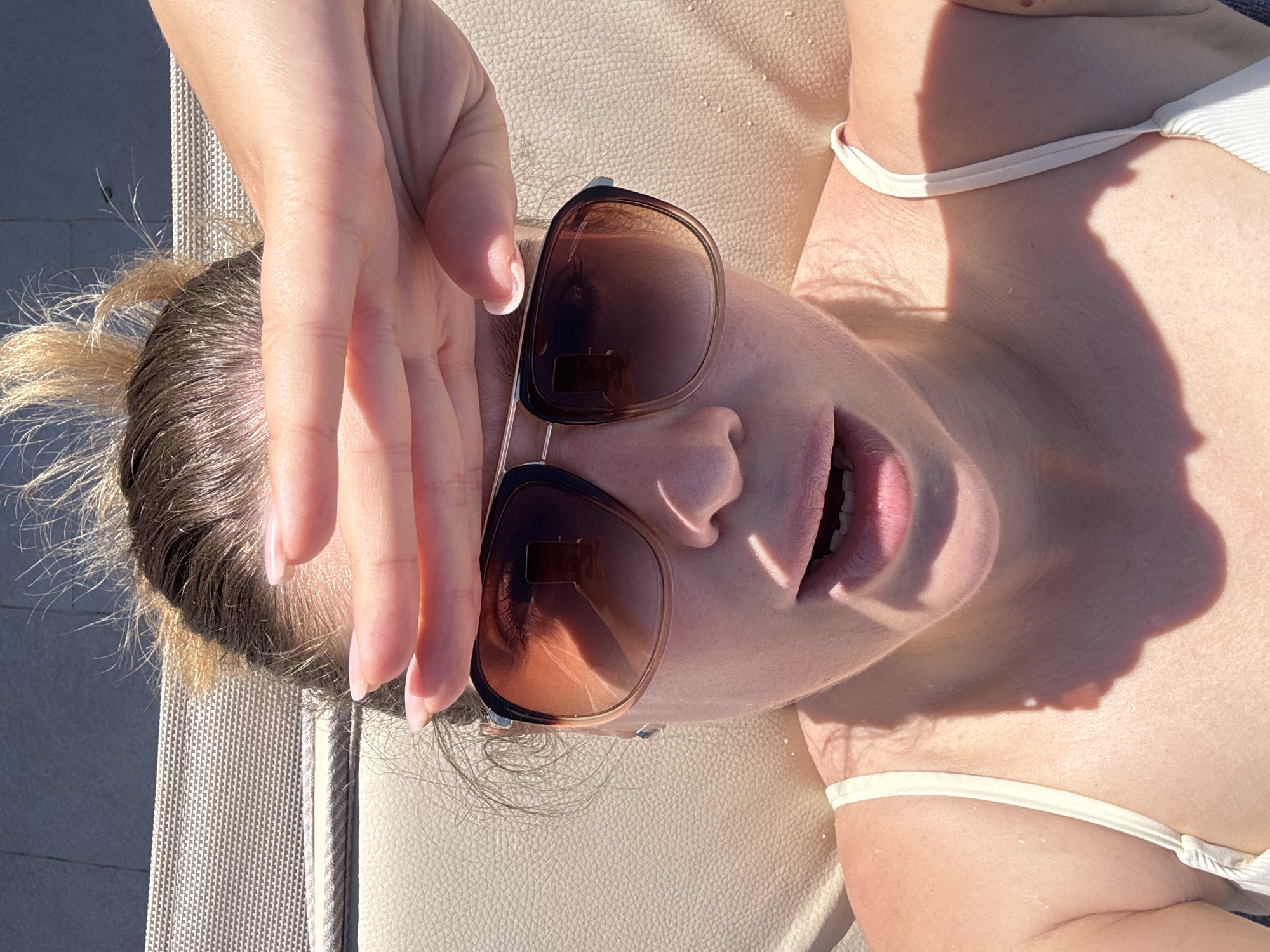 A woman is lying on a beige lounge chair outdoors. She is wearing a white bikini top and sunglasses. She has her hand raised near her face. The background includes textured beige material of the lounge chair and hints of an outdoor environment. The location is likely Đenovići, Montenegro.