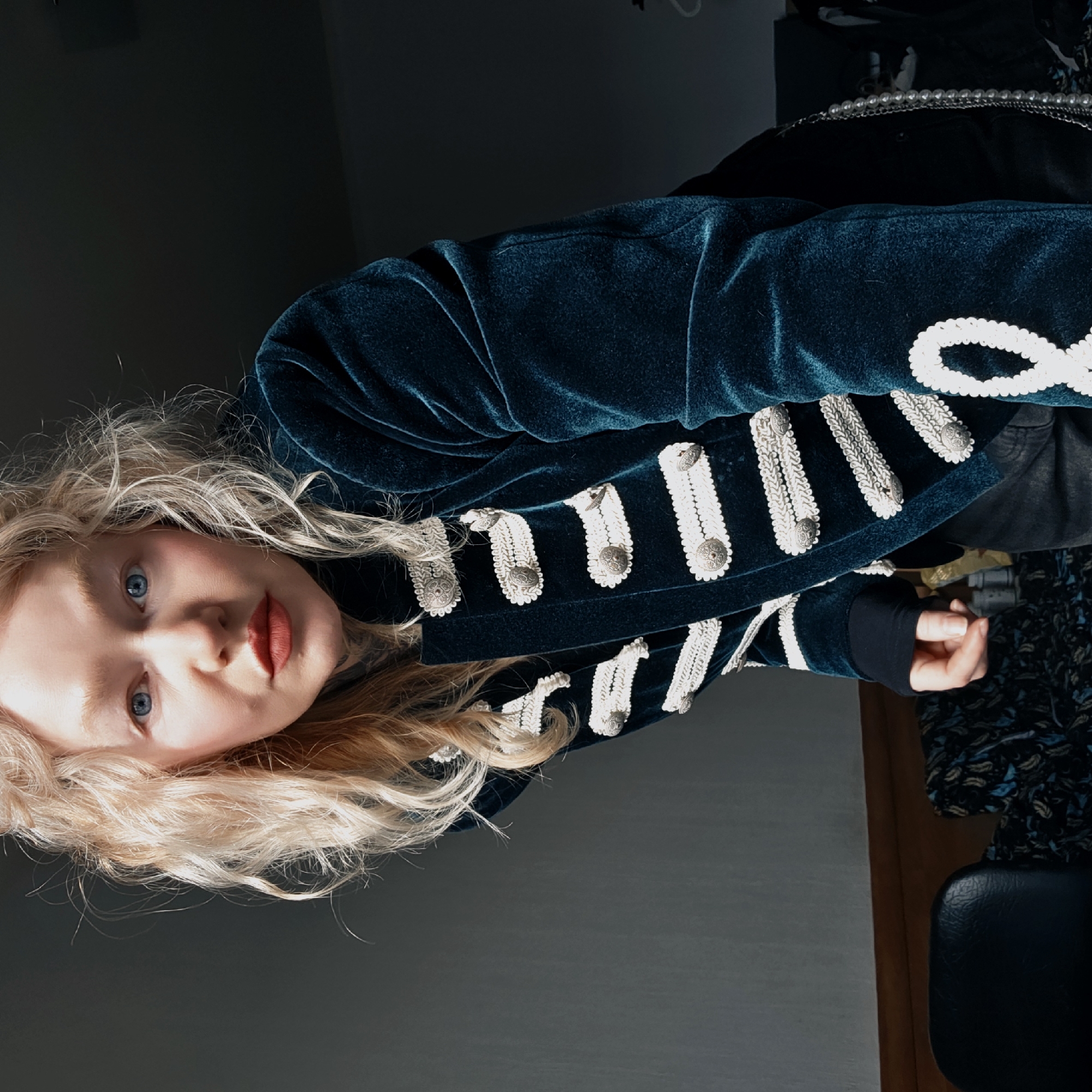 A fair-skinned individual with long, blonde, curly hair and blue eyes is wearing a teal velvet jacket with white decorative trim and silver buttons. The person is wearing red lipstick and looking directly at the viewer. The background is a dimly lit room with grey walls. A black chair and patterned clothing are visible in the background to the right of the individual. The image does not provide any clues about the location.