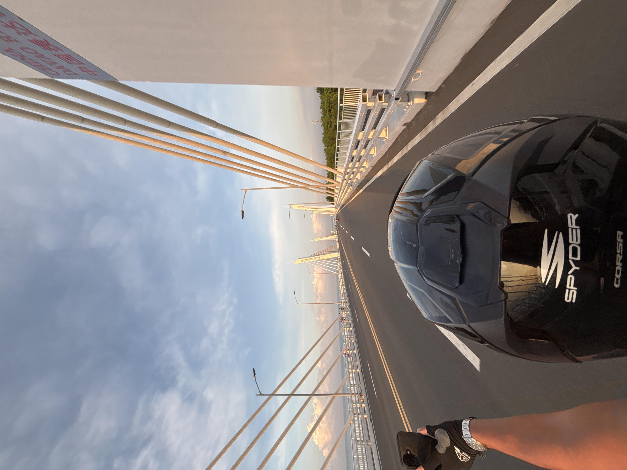 The image shows a first-person perspective of someone riding a motorcycle on the Viaduct in Davao City, Philippines. The rider is wearing a black helmet with "SPYDER CORSA" written on it. They are holding up a smartphone in their left hand, presumably to take a photo or video. The rider is on a multi-lane paved road on a cable-stayed bridge. The sky is blue with some scattered white clouds, and the sun appears to be setting, casting a warm glow on the bridge's cables and the surrounding environment. In the distance, the bridge extends towards the horizon, with other similar bridge structures visible. On the right side of the image, a large white concrete structure with text in Chinese and English is visible. The text on the structure reads "CHINA AID DAVAO H...". On the left side of the road, there is a railing and then a view of the ocean with some pinkish clouds. There are a few other people visible on the bridge walkway, and some vegetation can be seen in the far distance. The overall scene suggests a clear day with pleasant weather, and the rider is enjoying a scenic journey across the bridge.