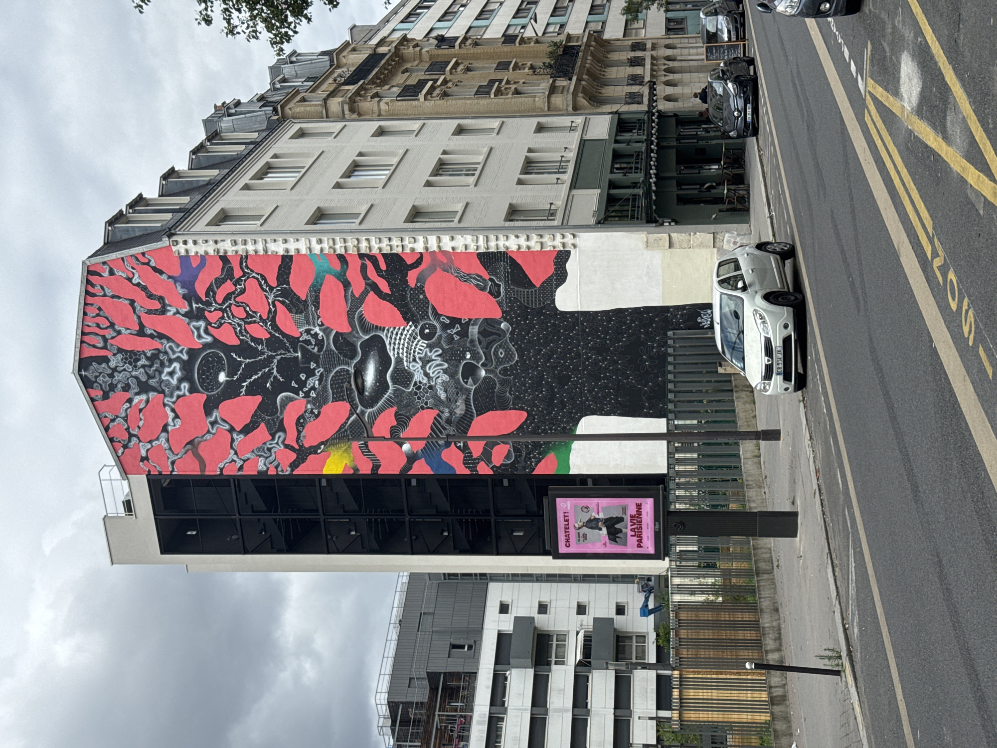 An urban street scene is depicted under an overcast sky during daytime. The central feature is a large, vibrant mural covering the side of a multi-story building. The mural is primarily black and red, with intricate white and grey patterns. Large, irregular red shapes resembling organic forms or leaves are interspersed with a textured black background filled with detailed, abstract designs. Hints of blue, yellow, and green paint are also visible within the mural. Below the mural, the building features modern dark glass windows.
To the right of the muralled building, an older, more ornate building with classic Parisian architecture, including a mansard roof and detailed cornices, stands tall. Further to the right, a street with a curb, sidewalk, and road is visible. A white Dacia Sandero car is parked on the street next to the sidewalk, and another dark car is parked further down the street. Yellow lines on the asphalt indicate road markings, including a partially visible "BUS" inscription, suggesting a bus lane or stop.
On the sidewalk near the mural, a digital billboard displays a pink advertisement. Visible text on the billboard reads "CHATELET!", "LA VIE PARISIENNE", "OFFENBACH", and "OPÉRA", along with "Gyps". In the background, modern buildings with lighter facades and darker window frames are present. Trees are partially visible at the top of the image against the cloudy sky.