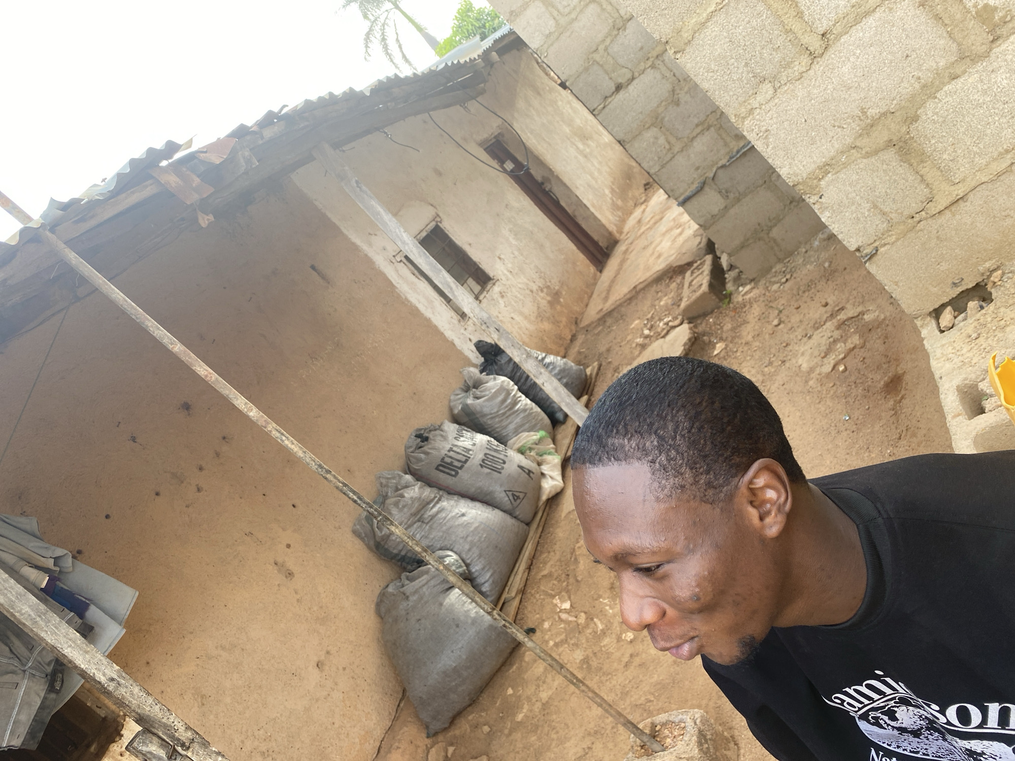 This image captures an outdoor scene in Bukuru, Nigeria, from a slightly elevated and tilted perspective. The overall environment appears functional, featuring rustic construction and earthy tones under bright daylight.
In the foreground, partially visible on the right, is a man with dark, closely cropped hair and brown skin. He is wearing a black long-sleeved shirt with some white printing that partially reads "...ami...son". The man is looking down and to his right, displaying a neutral or pensive expression.
The background is dominated by a structure with textured, possibly mud or earthen walls, topped by a corrugated metal roof that shows signs of wear. Along the base of this wall, a row of large, grey-white sacks is stacked. Visible text on one sack indicates "DELTA SM..." and "100 KG", accompanied by a hazard symbol, suggesting they contain some material or product. Several raw wooden or metal poles crisscross the scene, appearing to be structural supports for the roof or part of a simple enclosure.
To the far right, a wall made of concrete blocks is visible, contrasting with the more organic texture of the main building. The ground is bare, reddish-brown earth, indicative of the local soil. In the upper background, the bright, possibly overcast sky and the fronds of a palm tree are visible, confirming the outdoor, daytime setting. Two small windows and a doorway are discernible on the distant wall of the main structure. The scene conveys a sense of a practical, possibly working or storage area.