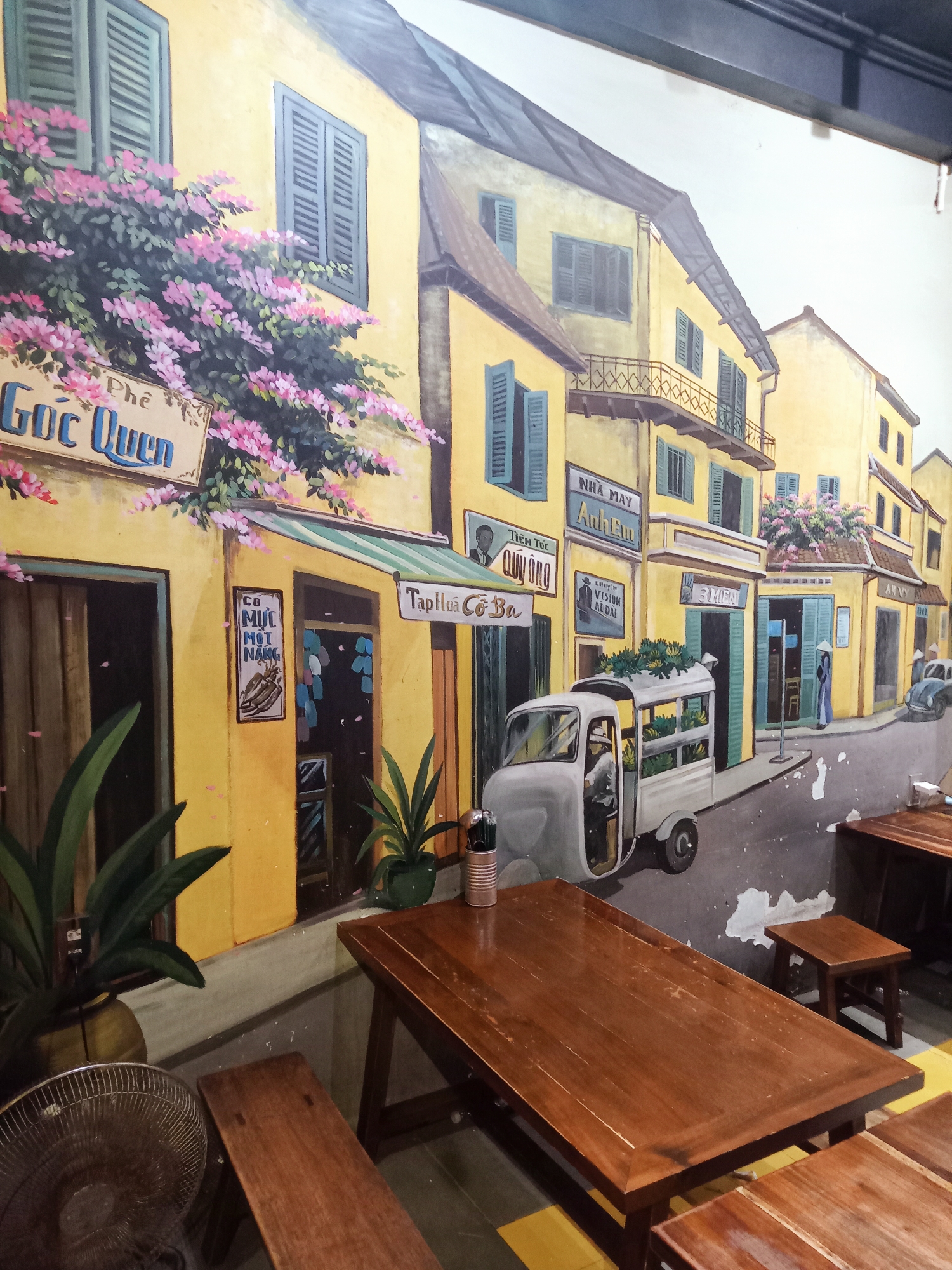 The image displays an indoor wall covered by a detailed mural depicting a charming street scene in a Vietnamese town, characteristic of locations like Nha Trang. The mural features two-story yellow buildings with blue and green shuttered windows, some adorned with cascading pink bougainvillea.
On the left, a sign reads "Phê Góc Quen" (Coffee Familiar Corner), with another below it saying "CƠ MỰC MỘT NẮNG" (Sun-dried squid). Other visible shop signs include "TIỆM TÓC QUÝ ÔNG" (Gentleman's Barbershop), "Tạp Hóa Cô Ba" (Ms. Ba's General Store), "NHÀ MAY Anh Em" (Anh Em Tailor Shop), "CHUYÊN VISION AL ĐẠI" (specializing in eyewear), "3 MIỀN" (3 Regions), and "AN VY".
A white, three-wheeled utility vehicle, possibly a vendor's cart, is parked on the street, laden with green produce like bananas, and a person is visible in the driver's seat. Further down the street, a figure wearing a traditional conical hat (nón lá) and áo dài stands near a doorway, and a vintage car is parked. The scene is set during the daytime, indicated by the bright light.
In the foreground, real dark wooden tables and benches are arranged for customers, blending with the painted environment. A metallic fan is visible on the left, and actual potted plants are placed near the mural, enhancing the immersive effect of the depicted street. The overall impression is that of a cozy cafe or restaurant with a nostalgic, artistic ambiance.