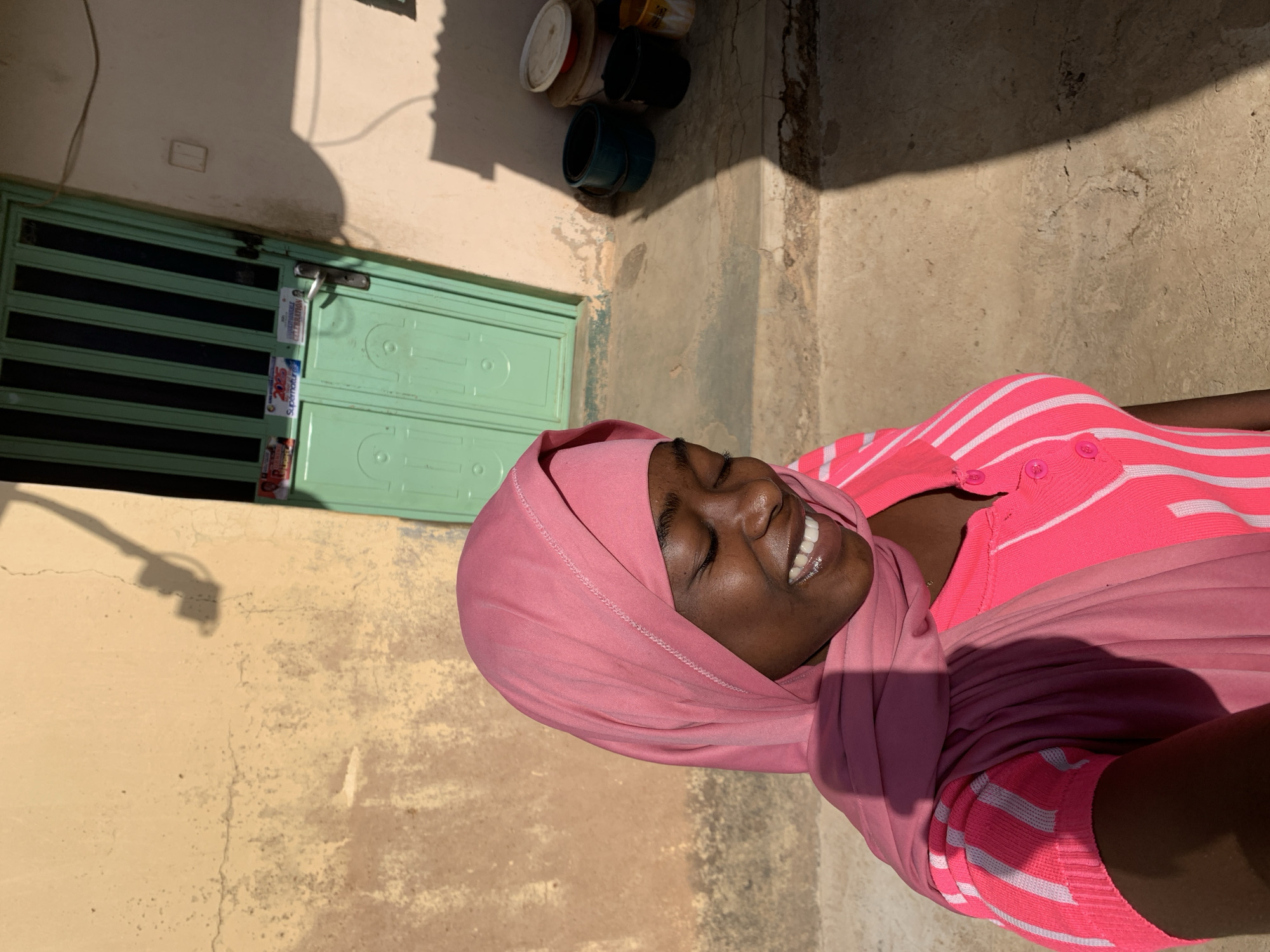 A medium shot captures a smiling dark-skinned woman wearing a pink hijab and a pink and white striped cardigan. The scene appears to be outdoors, next to a light yellow wall and a green door with horizontal black bars. Above the door are stacked buckets and containers. The ground is a light-colored concrete. A shadow from a street light is cast on the wall next to the door. The location is Rafin Par, Nigeria.