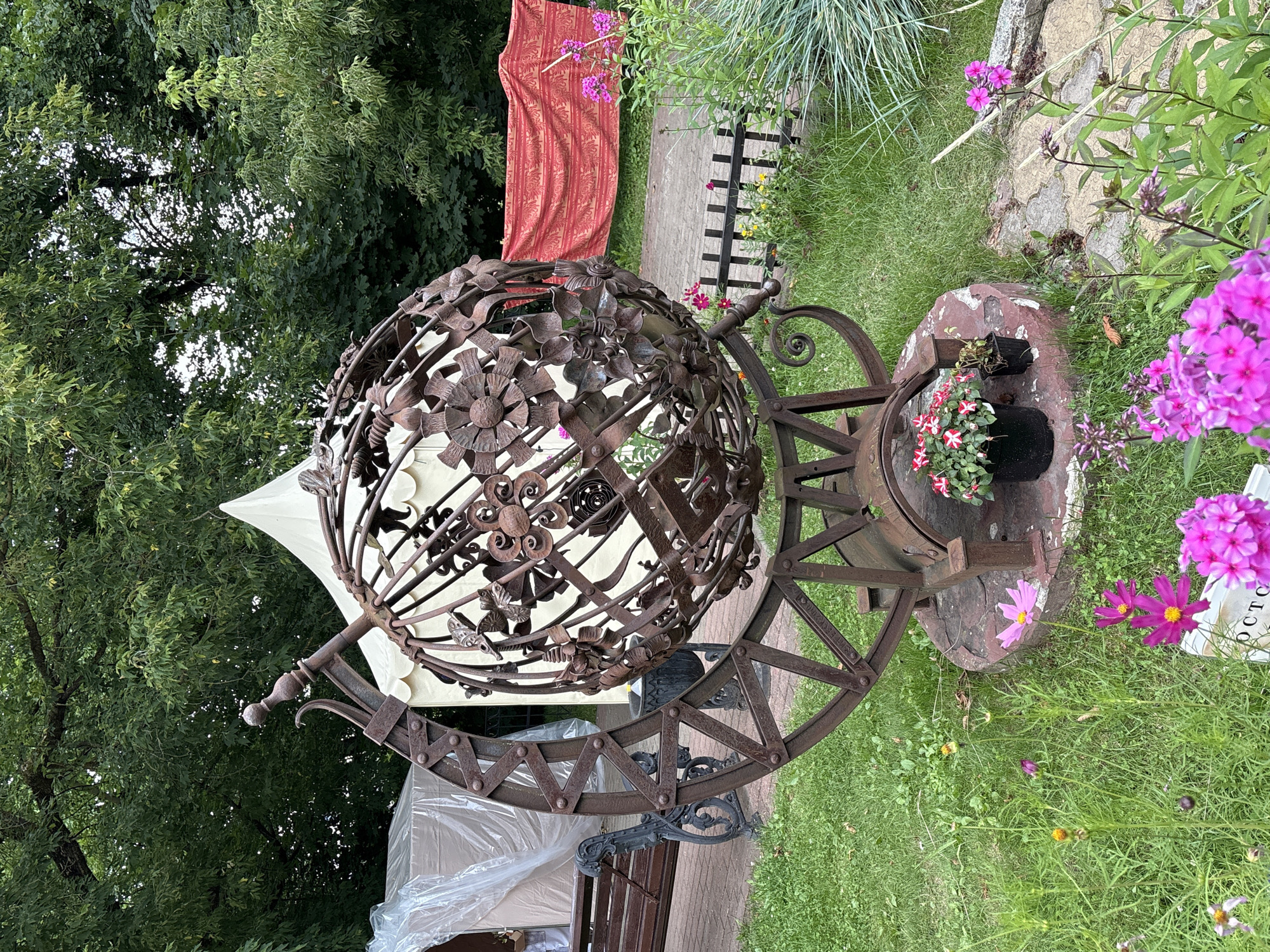 The image shows a large, rusted metal globe-like sculpture in a park-like setting. The sculpture is ornate, made of intricate metalwork that resembles flowers and leaves. It is supported by a circular metal base. In the background, there are trees, a white tent, and a red patterned fabric hanging on a fence. There are also flowers in pots near the base of the sculpture, and pink flowers are blooming in the foreground. A wooden bench and a covered structure are visible to the left. The ground is covered in grass and a stone pathway.
The sculpture is a prominent feature in the Museum of Vitebsk History, located in Vitebsk, Belarus. The sculpture is identified as "The Globe" and was created by the Vitebsk artist Lev Melamed.