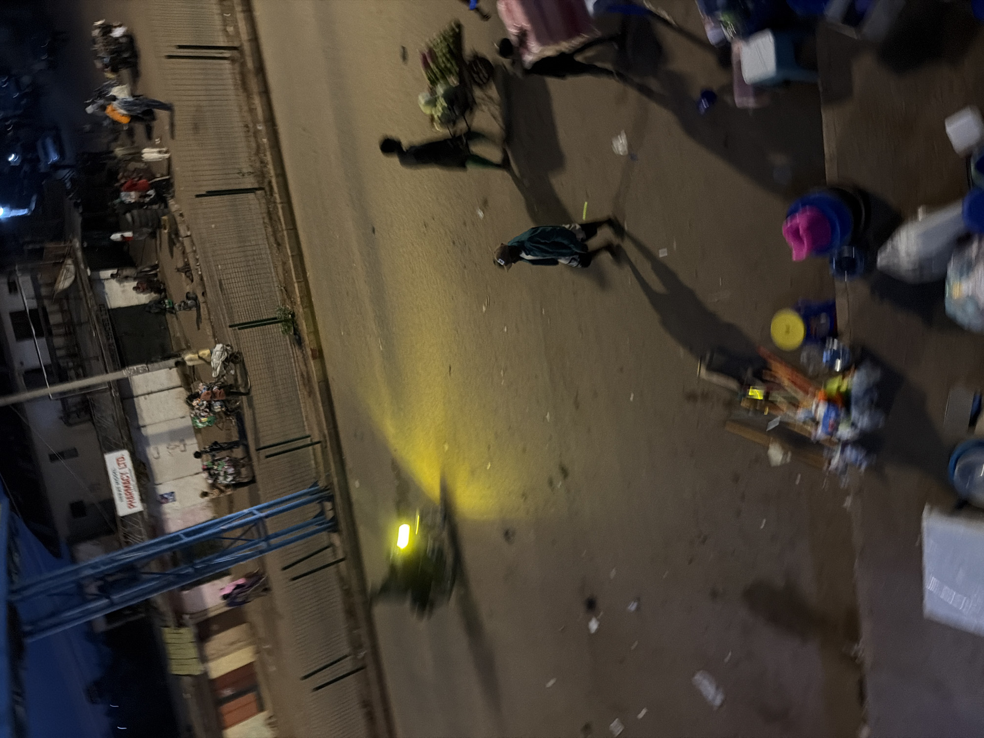 The image captures a dimly lit street scene in Bukuru, Nigeria, at dusk or night. A prominent blue metal structure, possibly part of a bus stop or shelter, dominates the left side of the frame. In the background, buildings line the street, with signage that includes "PHARMACY LTD." A street lamps casts a yellow glow on the asphalt road, illuminating several individuals and carts.
In the foreground, a person in a dark blue jacket and a baseball cap walks across the street, casting a long shadow. To their right, another person, dressed in dark clothing, pushes a cart laden with produce, including what appear to be watermelons and other vegetables. Further to the right, two other figures are visible, one carrying a large pink object and the other standing next to them.
The street is paved and shows signs of wear. Scattered across the road are pieces of litter, including plastic bags and wrappers. A few parked carts or stalls are visible along the sidewalk, some with goods displayed. The overall impression is of a bustling, commercial area at the end of the day, with activity continuing despite the fading light.