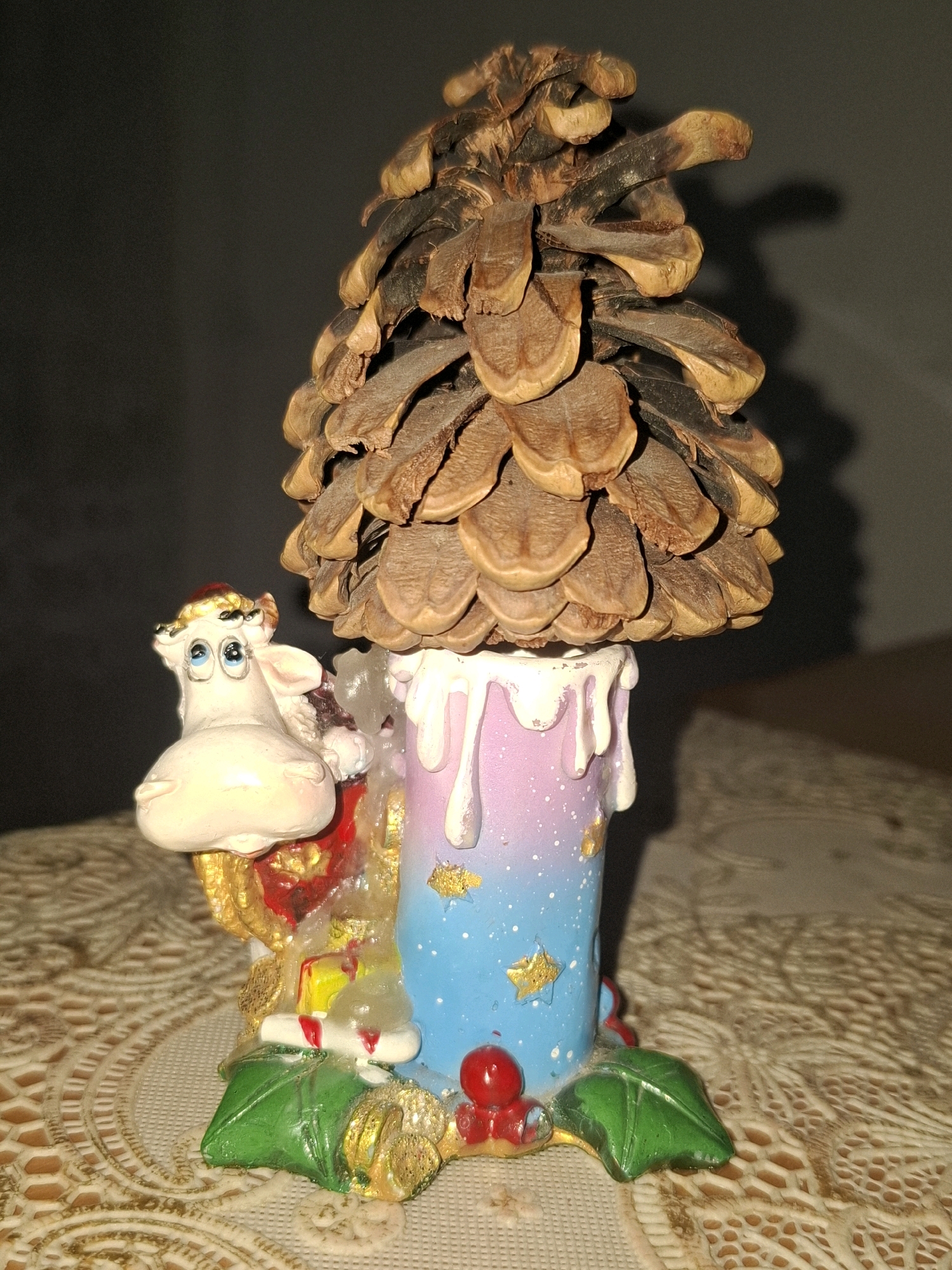 A close-up, eye-level, indoor shot features a decorative object resembling a pine cone atop a pastel-colored, candle-shaped base. The base is decorated with white dripping wax effects and scattered gold stars. To the left of the base is a small, whimsical figurine of a white cow with large blue eyes and a red and gold hat. The cow is adorned with gold accents and is positioned as if it is part of the candle's decoration. The object rests on a textured, beige surface with an intricate lace-like pattern. The background is dark and out of focus, with a hint of a wooden surface visible on the right. The lighting casts a shadow from the pine cone onto the dark background. The image does not provide any information about the city of Almaty, Kazakhstan, or any specific location within it.