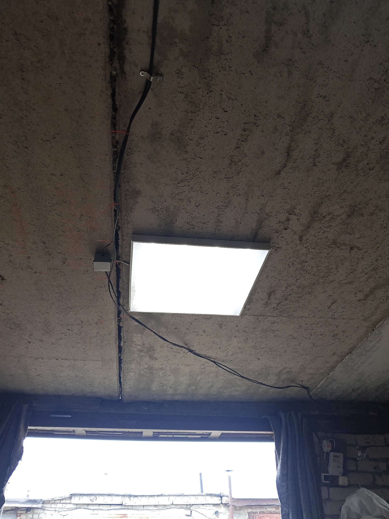 The image depicts a concrete ceiling in what appears to be an unfinished or industrial space. A rectangular LED panel light is installed, emitting a bright white light. Electrical wiring is visible, running along the concrete surface and connected to a junction box. The ceiling has a rough texture and a prominent vertical crack. Below the ceiling, a large opening, possibly a window or doorway, reveals an overcast sky and some brickwork and roofing in the distance. The overall scene suggests an indoor environment under construction or in a state of disrepair. No individuals are present in the image. The context of Chaikovsky, Russia, is not visually discernible from the provided image alone.