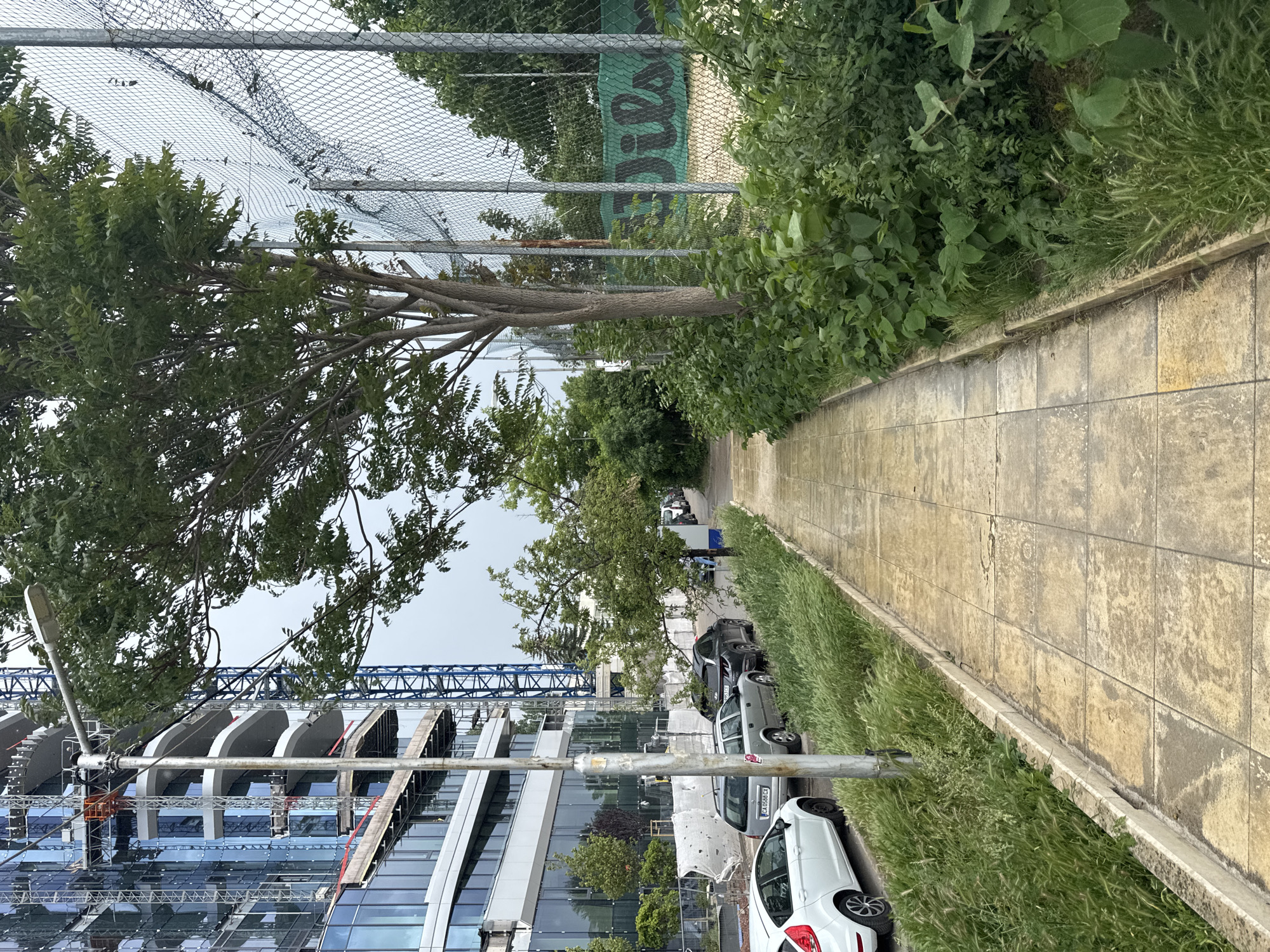 The image depicts a street scene in Sofia, Bulgaria. On the left is a modern building under construction, featuring a blue crane. A row of parked cars lines the right side of the street, next to a tiled pedestrian walkway. A chain-link fence runs along the left side of the walkway, with lush vegetation and trees adjacent to it. A sign with the word "Wilson" can be seen through the fence. The sky is overcast. There are some people visible on the sidewalk, though they're further off into the distance.