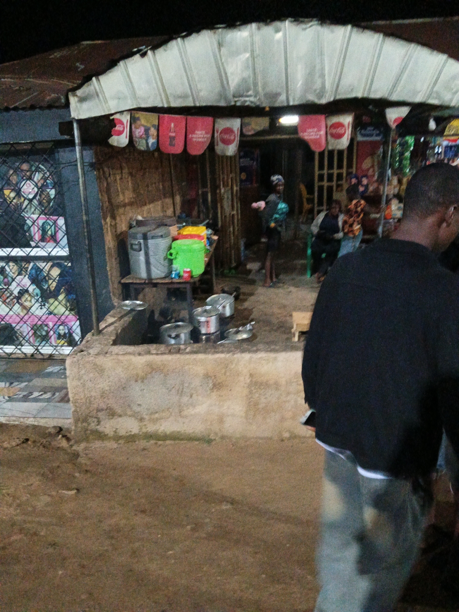 In Bukuru, Nigeria, an outdoor food stall is visible at night. A person in a black sweatshirt and jeans is in the foreground, partially obscuring the scene. The stall has a corrugated metal roof with white panels and several Coca-Cola branded banners hanging from it. In the background, several people are standing or sitting. A woman in a green top and headscarf is visible behind a cooking area with pots. Another person in a patterned top is also present. The ground appears to be dirt. The stall has a display of items behind a metal grate on the left side.