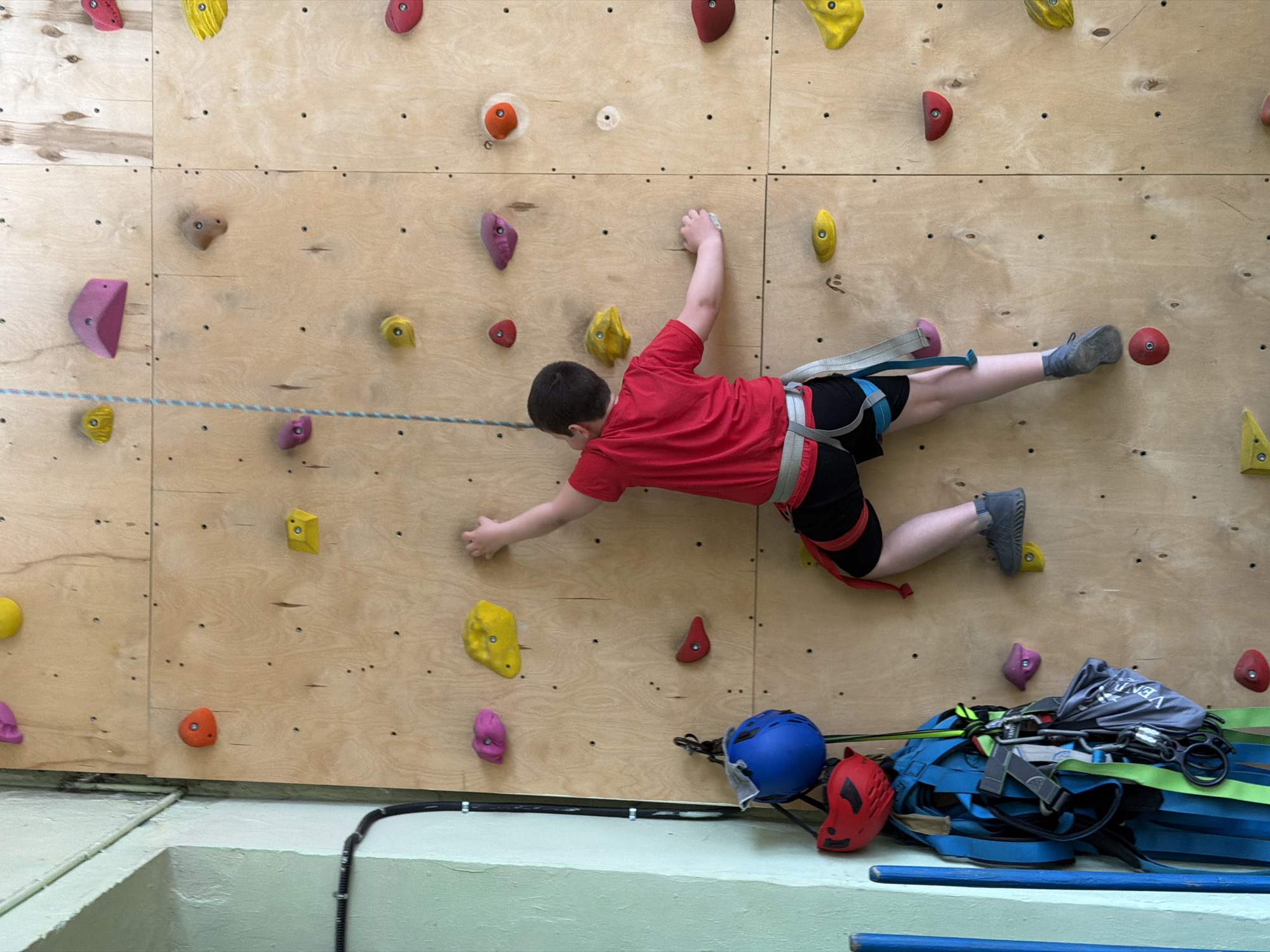 A child wearing climbing shoes, a red shirt, and black shorts is rock climbing on an indoor wall. The wall is constructed of wood panels and has various colored climbing holds attached to it. The child is wearing a harness. Climbing equipment, including helmets and ropes, is stacked on the ground below the climbing wall. The location is an indoor climbing facility. There is no specific evidence in the image to confirm the location in the city of Izhevsk, Russia.