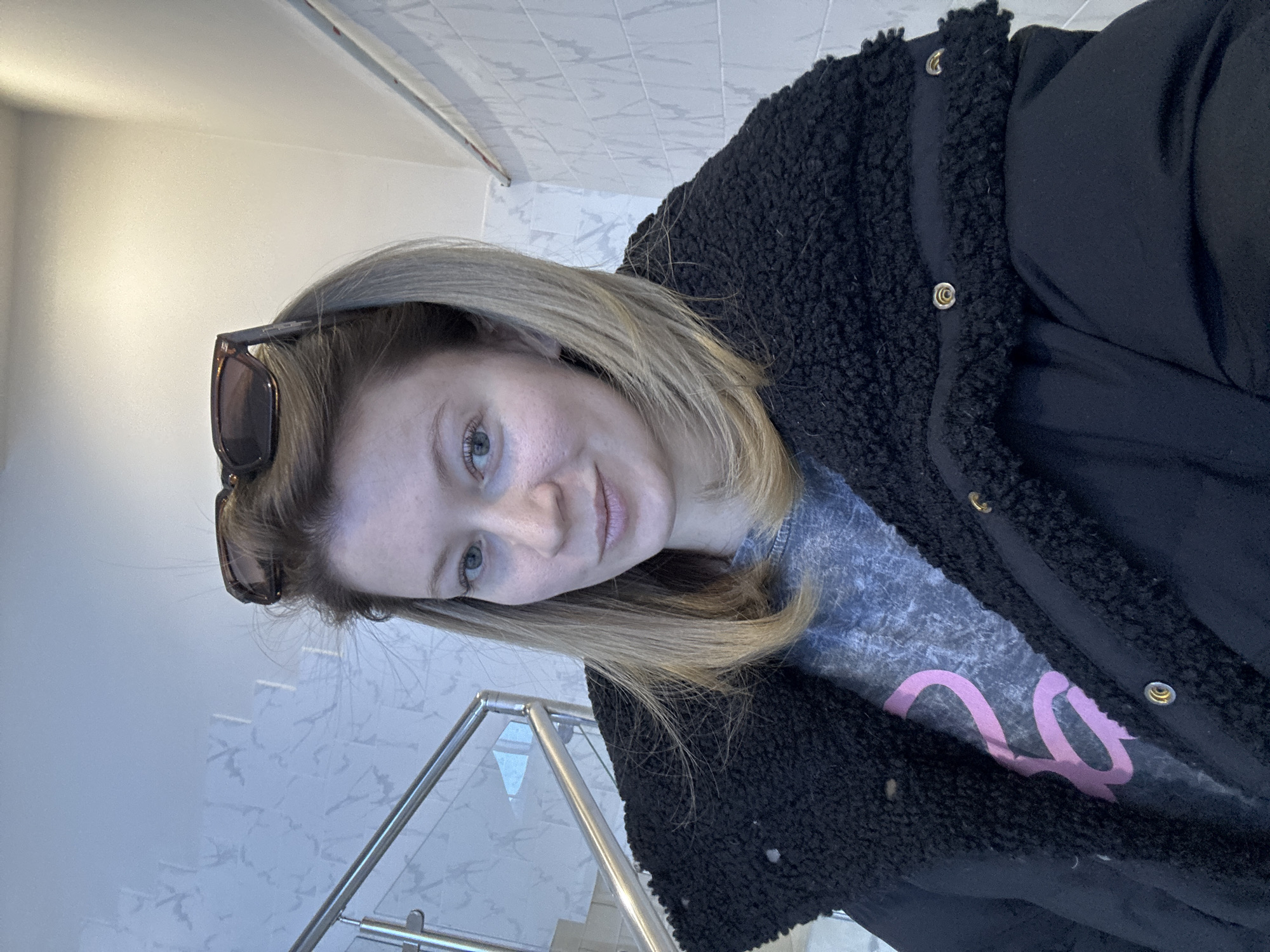 A fair-skinned female, estimated to be between 25 and 35 years old, with light brown hair and light-colored eyes, is centered in a close-up, chest-level portrait. She wears a black puffer jacket featuring a black, textured, faux-shearling collar and gold-toned snap buttons. A grey t-shirt with a partial pink graphic, possibly the numeral '2', is visible beneath the jacket. Tortoise-shell sunglasses are positioned on top of her head. The subject faces forward with a slight, closed-mouth expression. The background features a modern interior: a stainless steel railing with clear glass panels is visible on the left, indicative of a staircase or upper-level walkway. The wall to the left has a light grey patterned finish, while the wall on the right is covered in white, possibly ceramic, tiles with faint grey veining. The lighting appears even, suggesting an indoor environment. This scene is located within a building in the city of Chaikovsky, Russia.