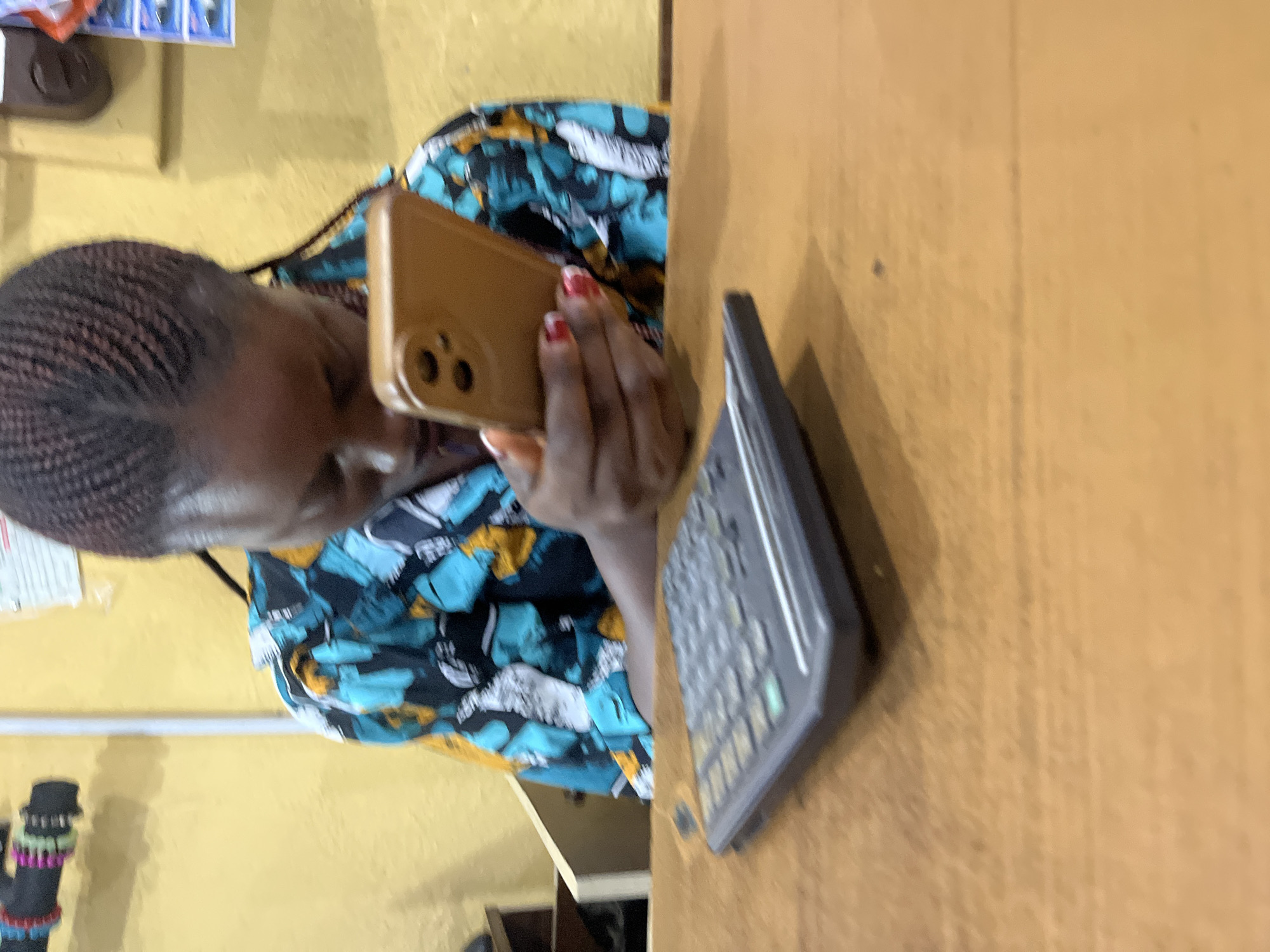 The media file displays a woman wearing a vibrantly patterned blue and yellow shirt, with her hair styled in braids. She is looking at a smartphone held in her right hand, which is adorned with red nail polish. To her right, on a wooden surface, is a black calculator. The background consists of a yellow wall, with some items visible on it, including what appears to be a calendar and some small boxes. In the lower left corner, a colorful object with black, pink, and green bands is partially visible. The scene is likely indoors, possibly in a shop or office, given the presence of the calculator and the general surroundings. The lighting suggests it is daytime. The image is oriented vertically, with the subjects appearing sideways.