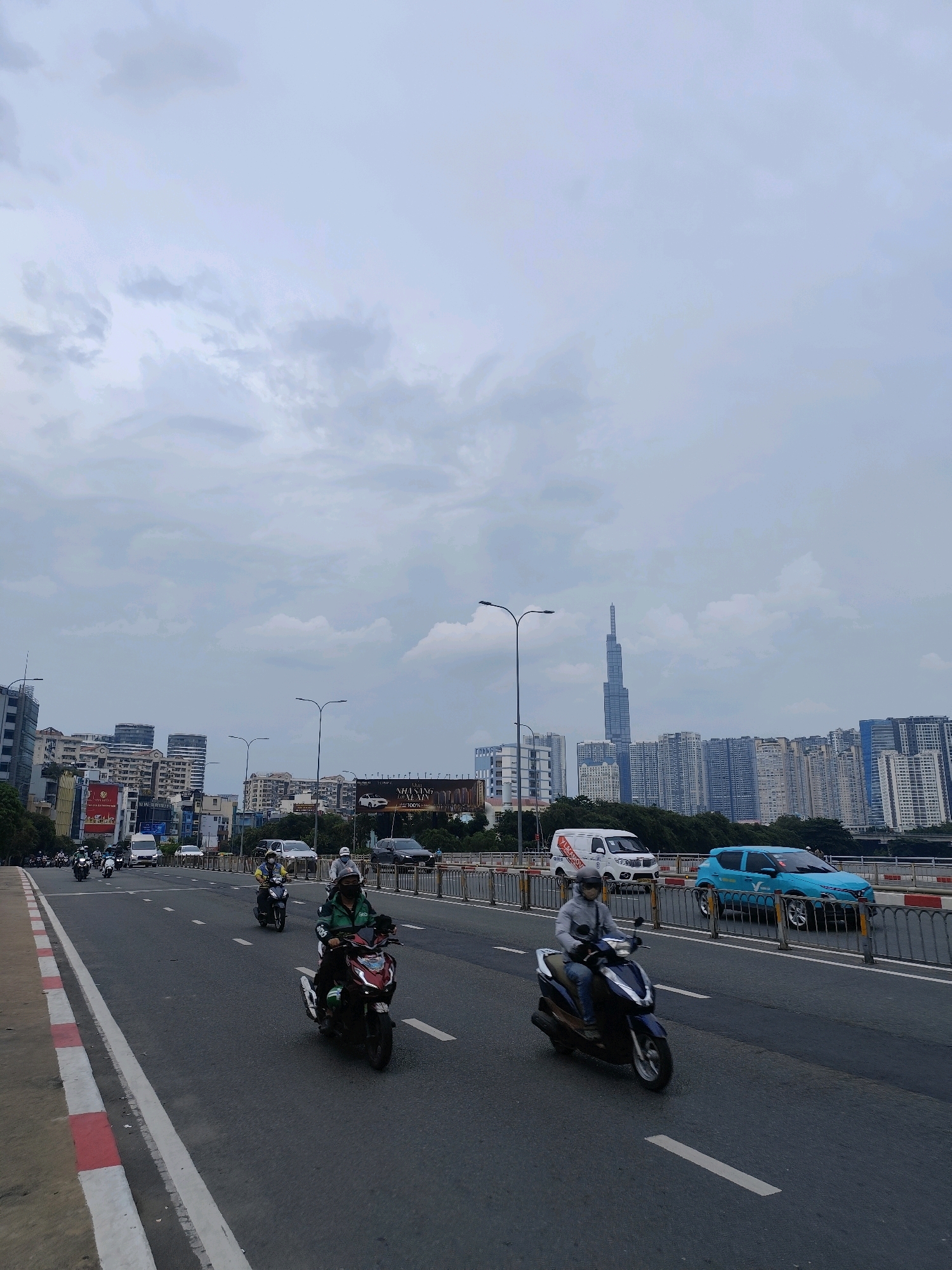 The image depicts a street scene in Ho Chi Minh City, Vietnam, likely on a bridge, with a cityscape in the background under a cloudy sky. The foreground features a multi-lane asphalt road with clear white lane markings. Several motorbikes are in motion. In the center-right, a rider wearing a green jacket and a face mask is on a dark red motorbike. To their left and slightly behind, another rider in a yellow shirt and black helmet is on a dark motorbike. Further ahead and to the right, a rider in a light grey jacket and blue jeans is on a dark blue motorbike. On the right side of the road, a turquoise car with a white logo is traveling. Beyond the road, a metal railing separates the roadway from a green embankment and trees. In the background, a dense urban skyline is visible, characterized by numerous high-rise buildings of varying architectural styles and heights. The most prominent structure is a very tall, slender skyscraper towards the left side of the background. Billboards are also visible on some of the buildings. The overall atmosphere is one of active city life on a slightly overcast day.