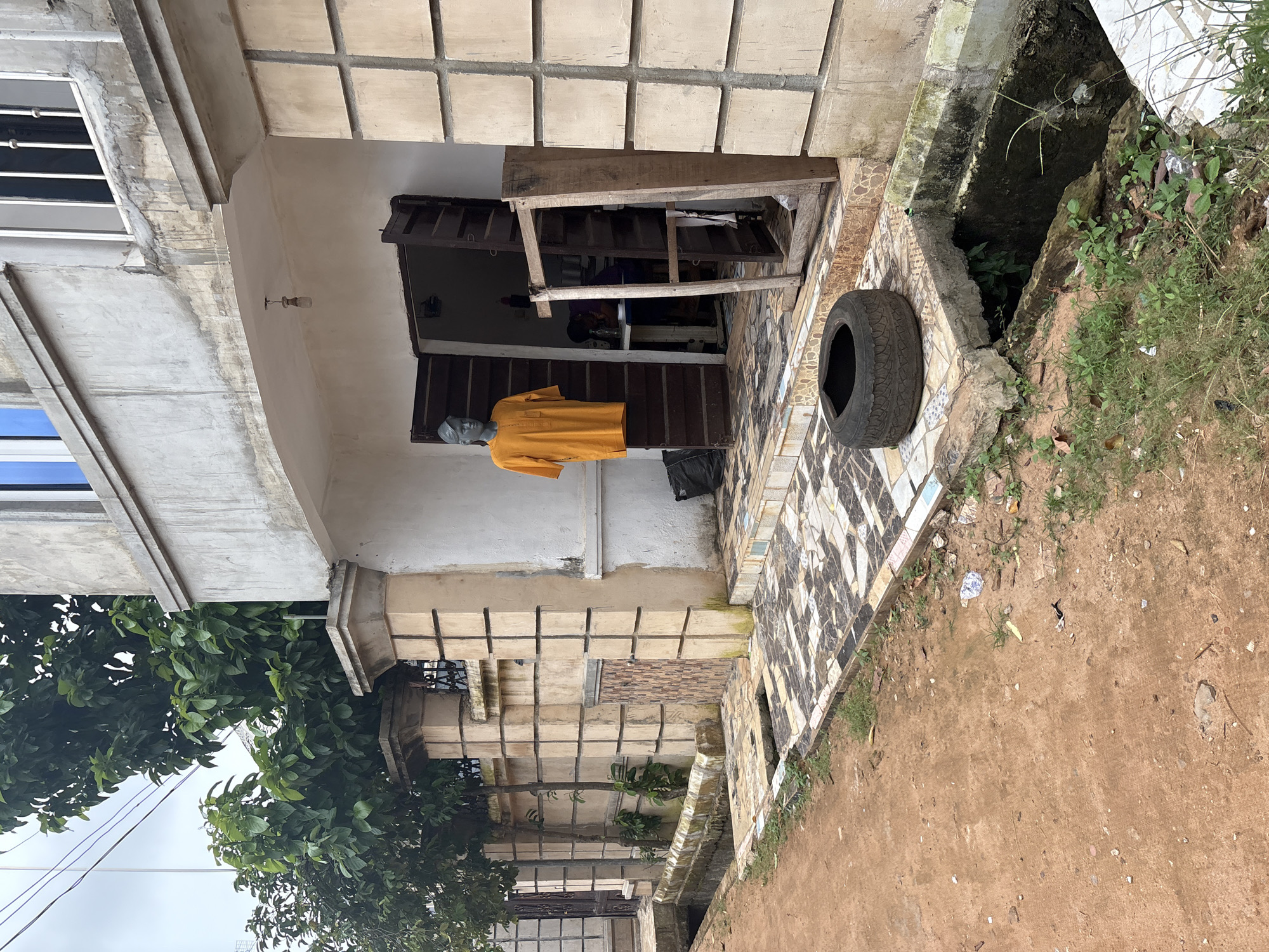 A low-angle, medium shot captures the facade of a shop in Obosi, Nigeria. The building, constructed with grey concrete and featuring beige tile accents, has an open entryway leading into a dimly lit interior. A mannequin stands just inside the doorway, displaying an orange garment. To the right of the entrance, a wooden frame leans against the wall. In front of the building, a weathered tire rests on a mosaic tiled path. A drainage ditch runs along the edge of the property, filled with murky water and overgrown vegetation. The ground in front of the shop is made of reddish-brown dirt, with scattered debris. Green trees and a stone wall with ornate metalwork flank the building on the left, adding to the scene's lived-in aesthetic.